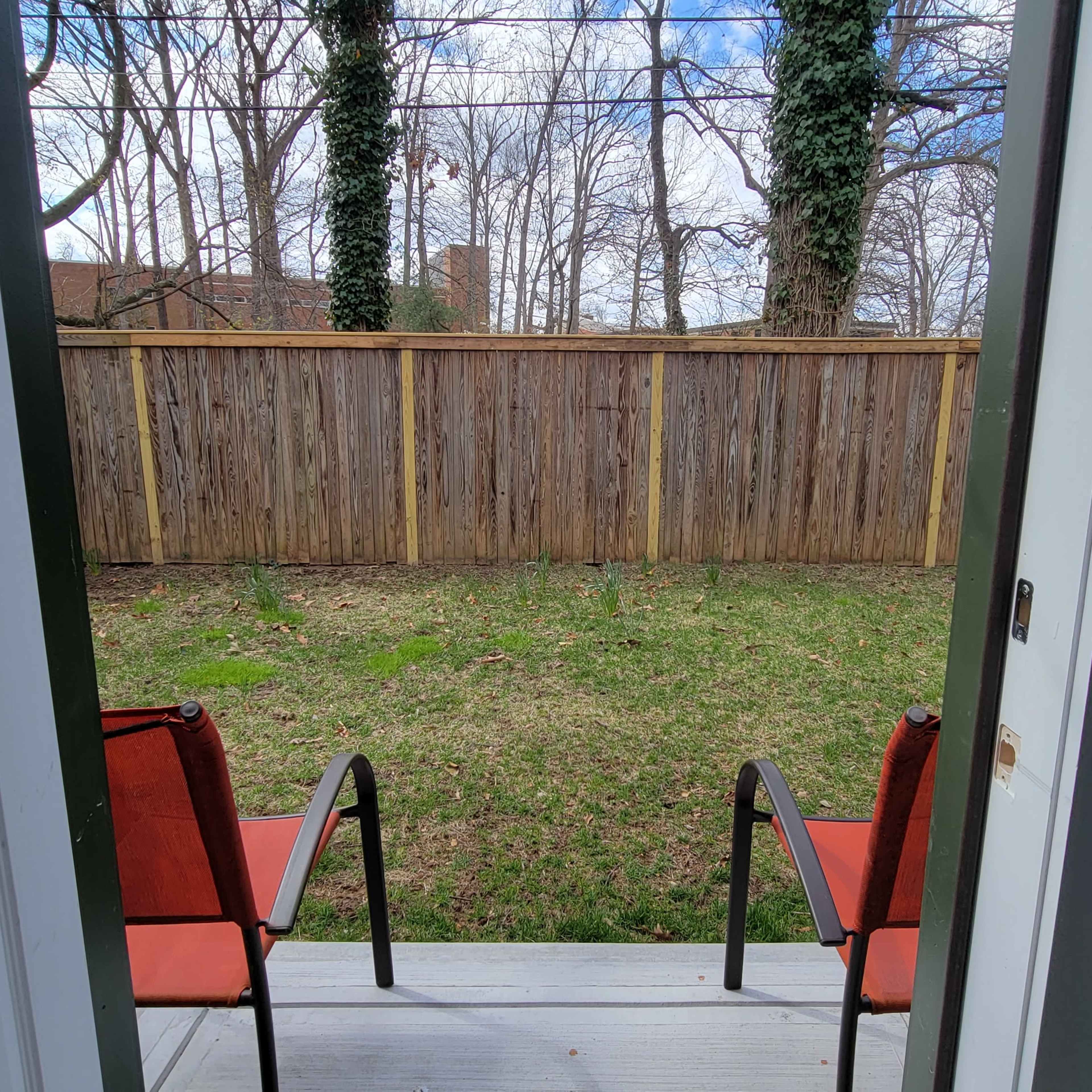 A view from an open door shows a small patio with two red chairs overlooking a grassy backyard enclosed by a wooden fence.