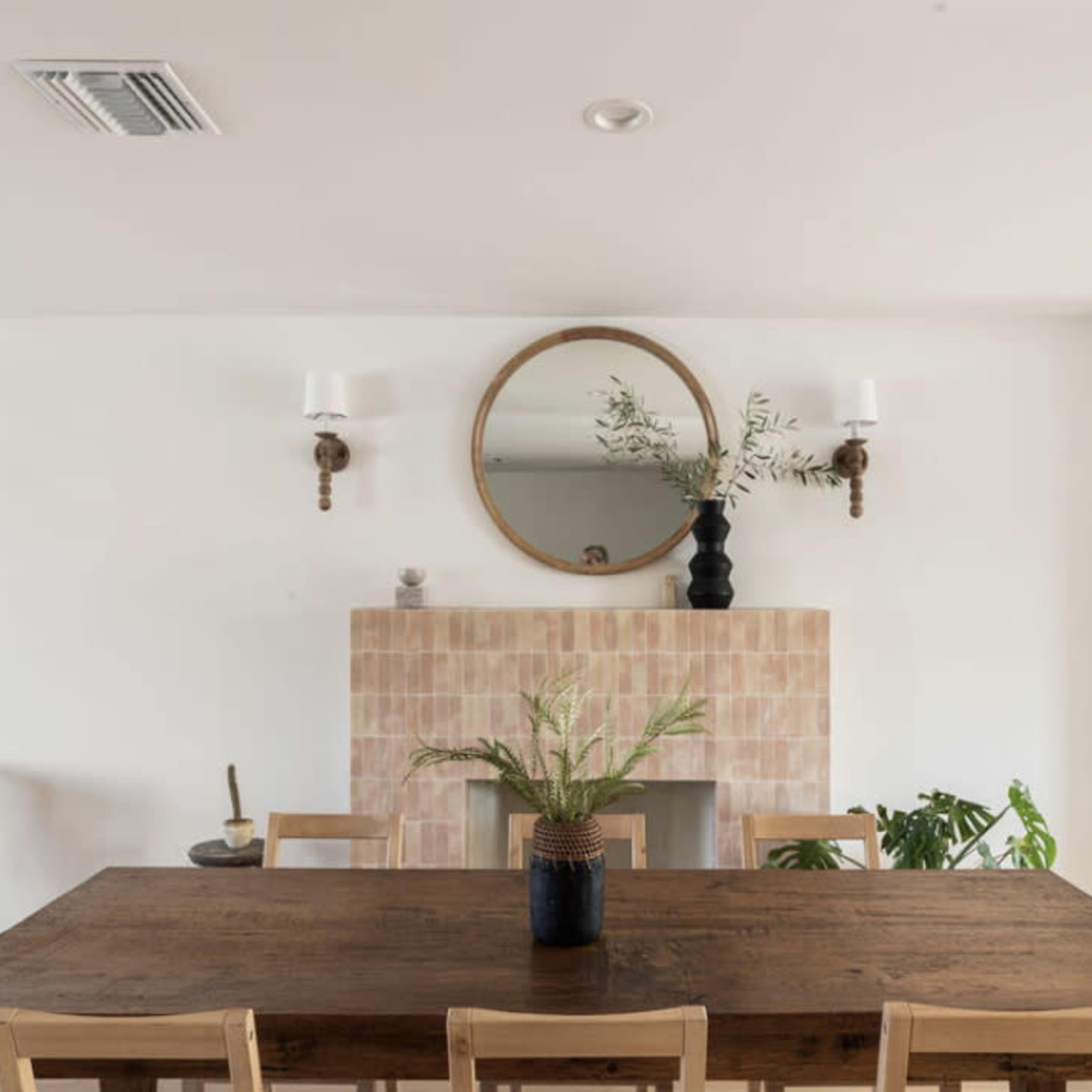 A modern dining area features a wooden table with light-colored chairs, a simple vase with plants, a round mirror above a tiled fireplace, and wall sconces.