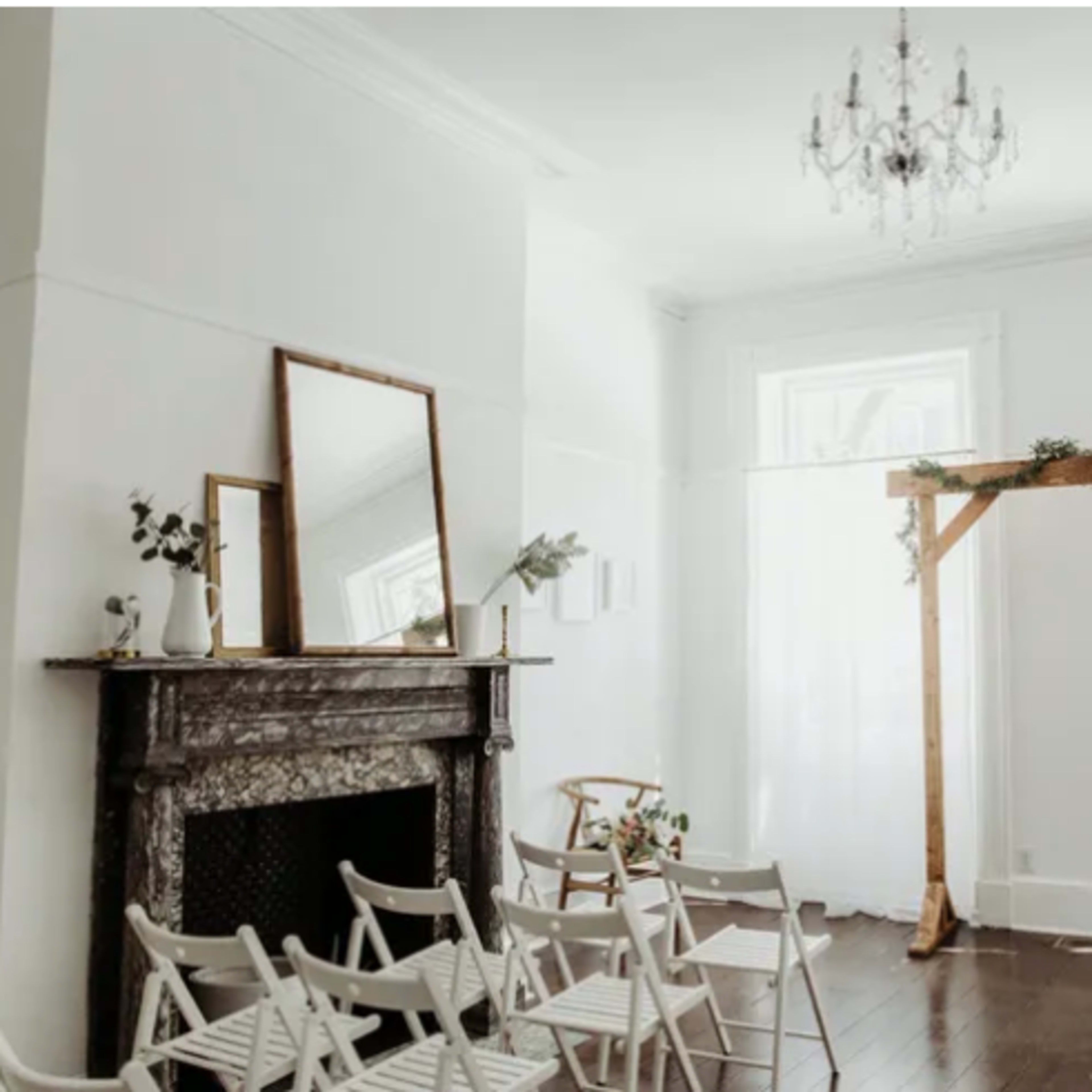 The room features a simple fireplace, a large mirror, and a wooden arch decorated with greenery, surrounded by white folding chairs.