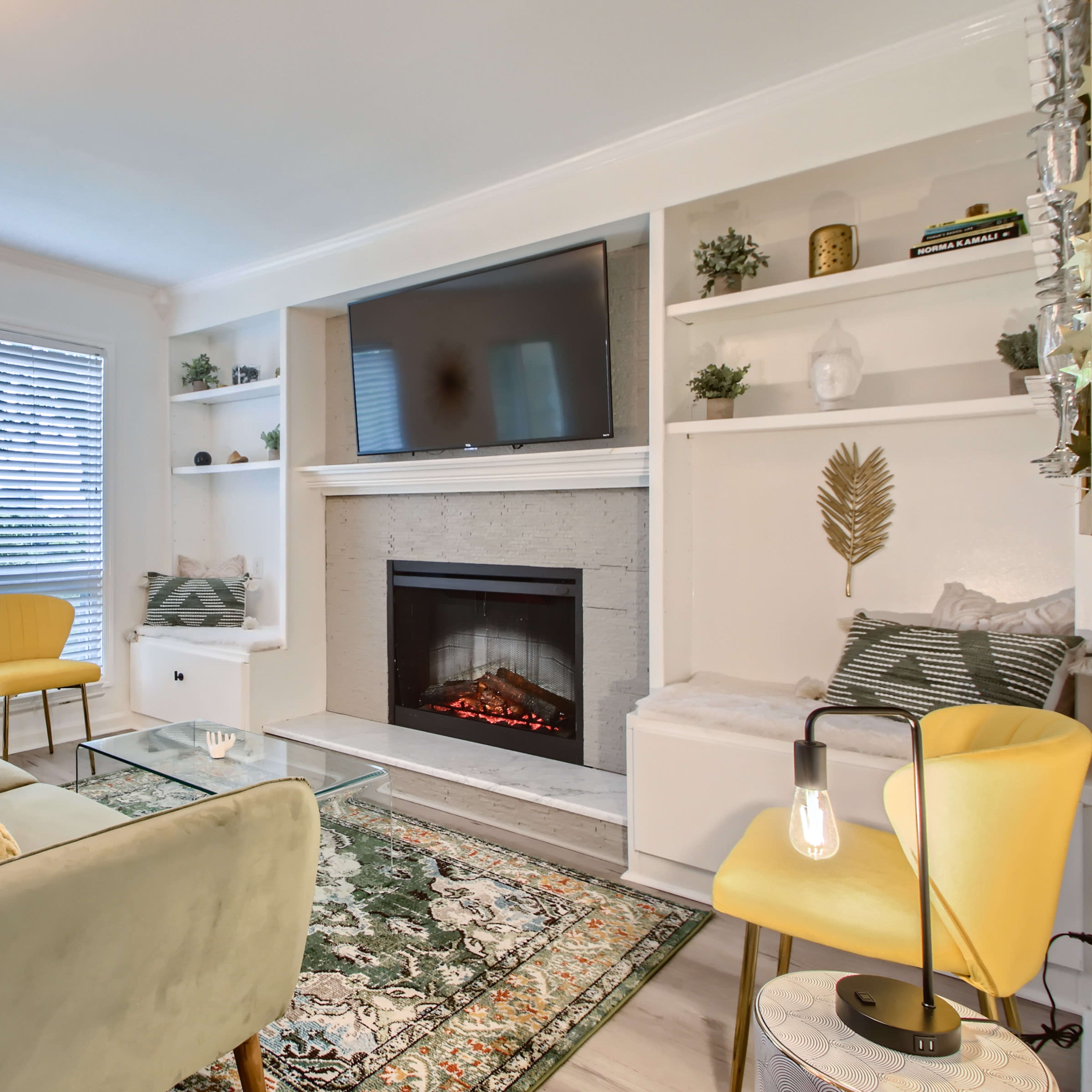 A modern living room featuring a cozy fireplace, a television above it, bookshelves on either side, and a pair of yellow chairs surrounded by a green sofa and a patterned area rug.