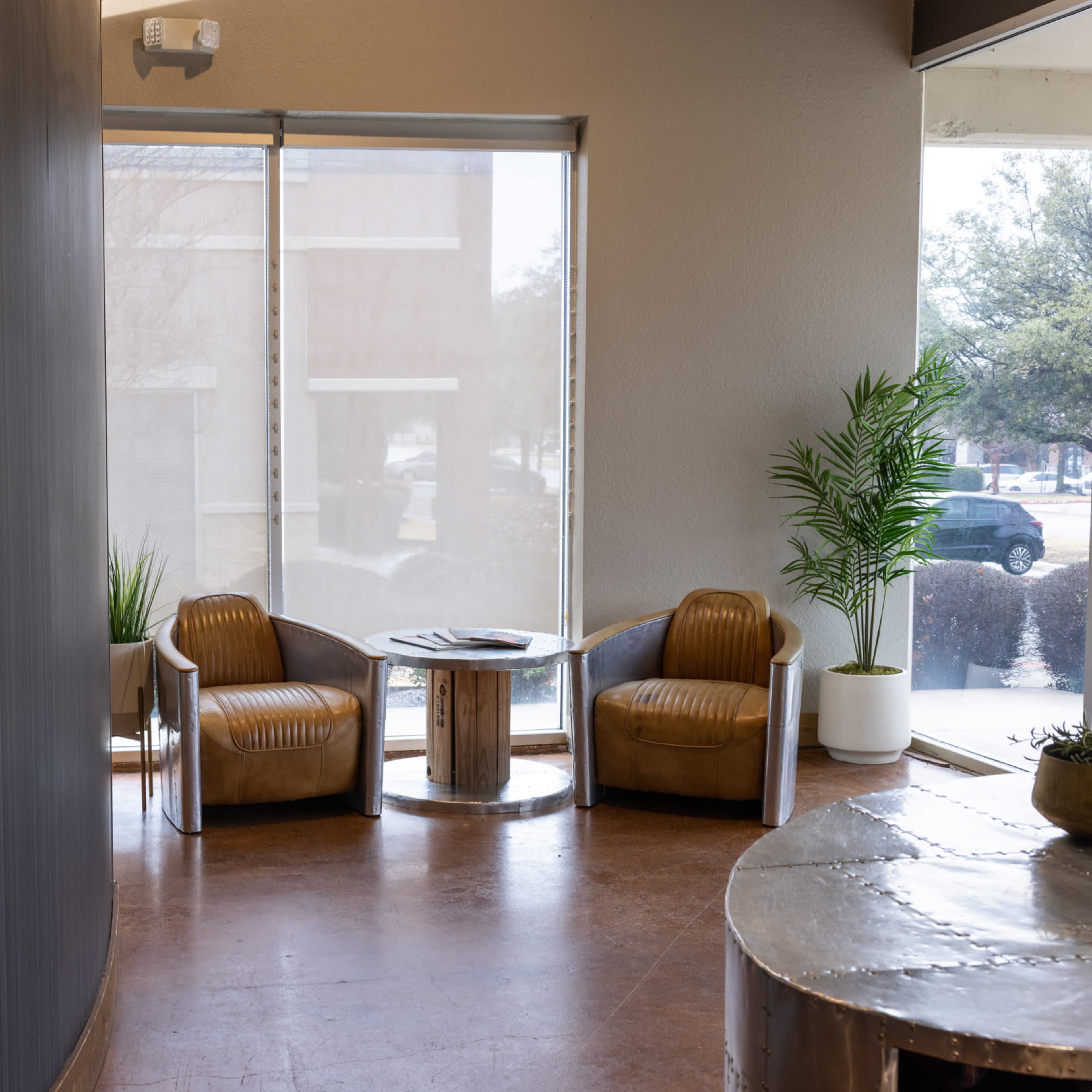 A modern waiting area featuring two brown chairs, a small table, and large windows letting in natural light.