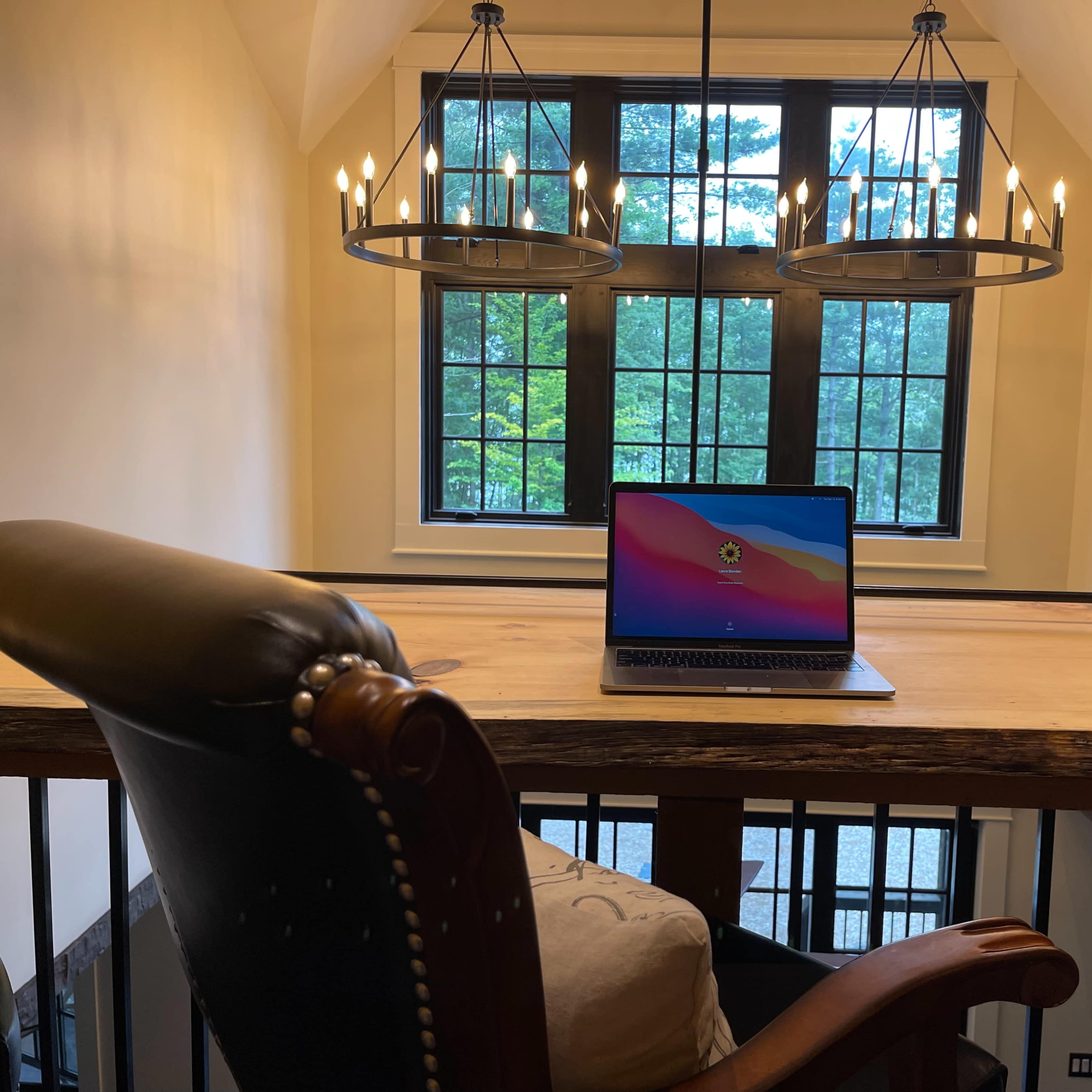 A laptop is placed on a wooden table beneath two circular chandeliers, with large windows in the background revealing greenery outside.