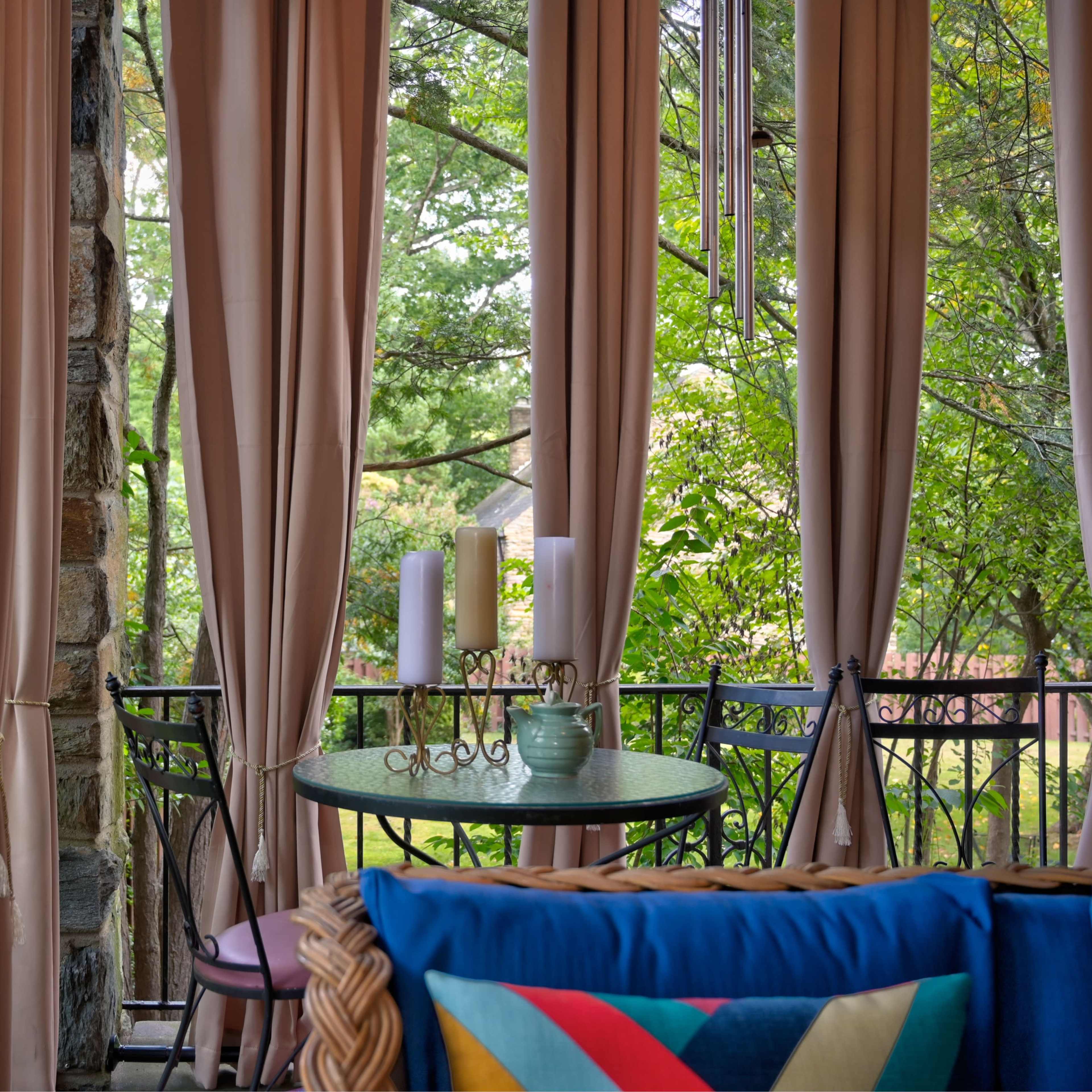 A balcony features a table and chairs, adorned with curtains and surrounded by greenery.