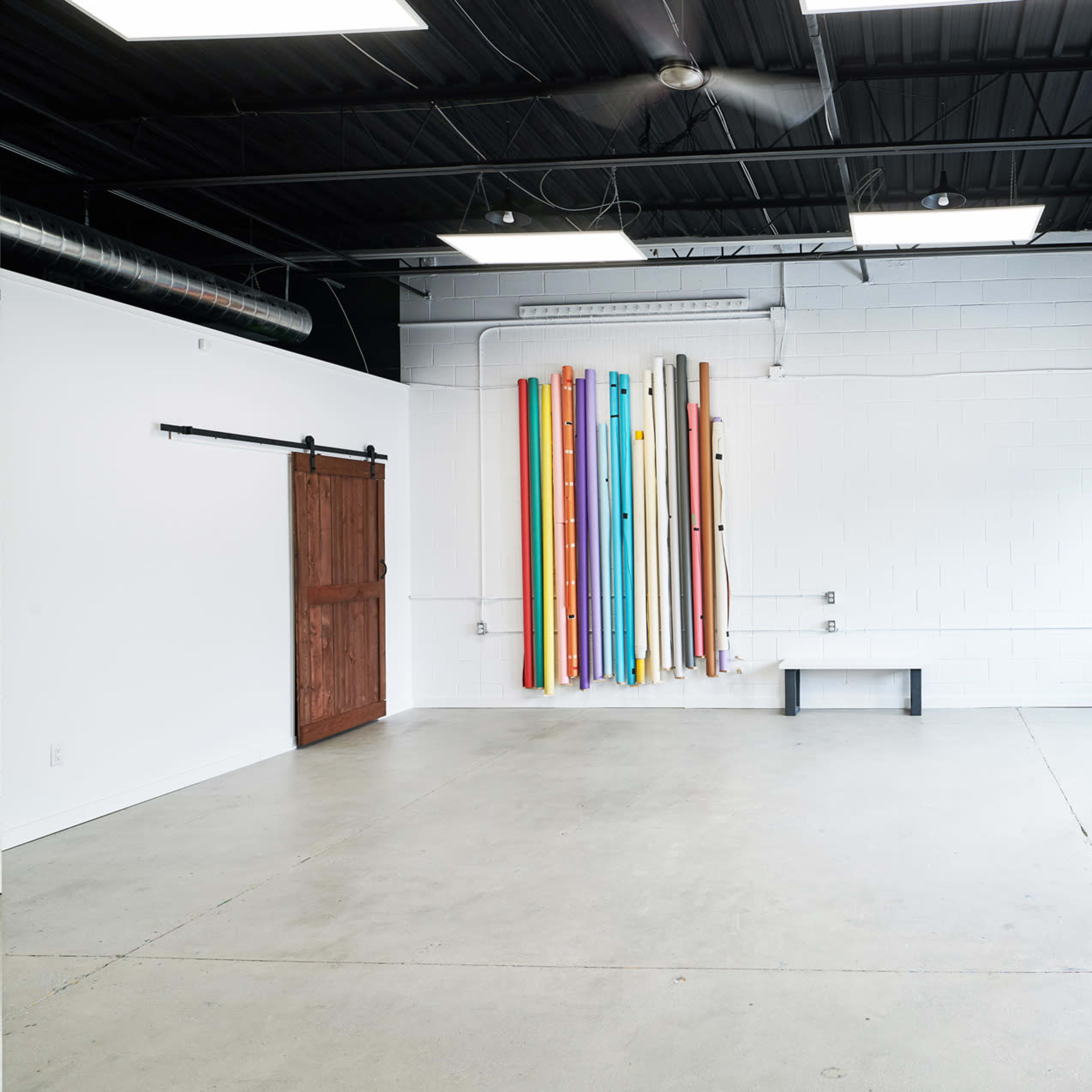 An empty room with a concrete floor, a wooden sliding door, colorful backdrops hanging on the wall, and large windows letting in natural light.
