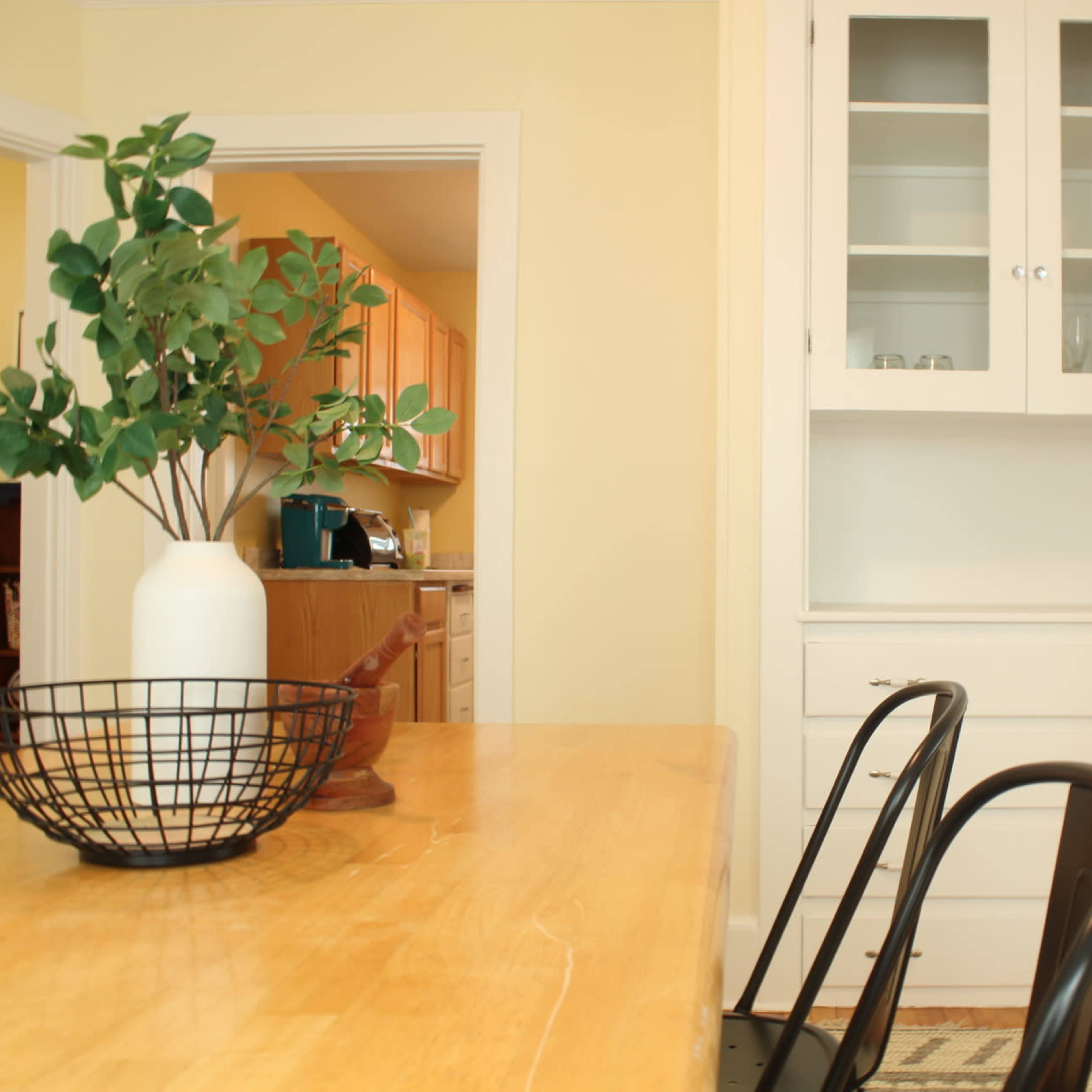 A light-colored wooden dining table with black chairs is positioned near a built-in cabinet in a bright kitchen area.