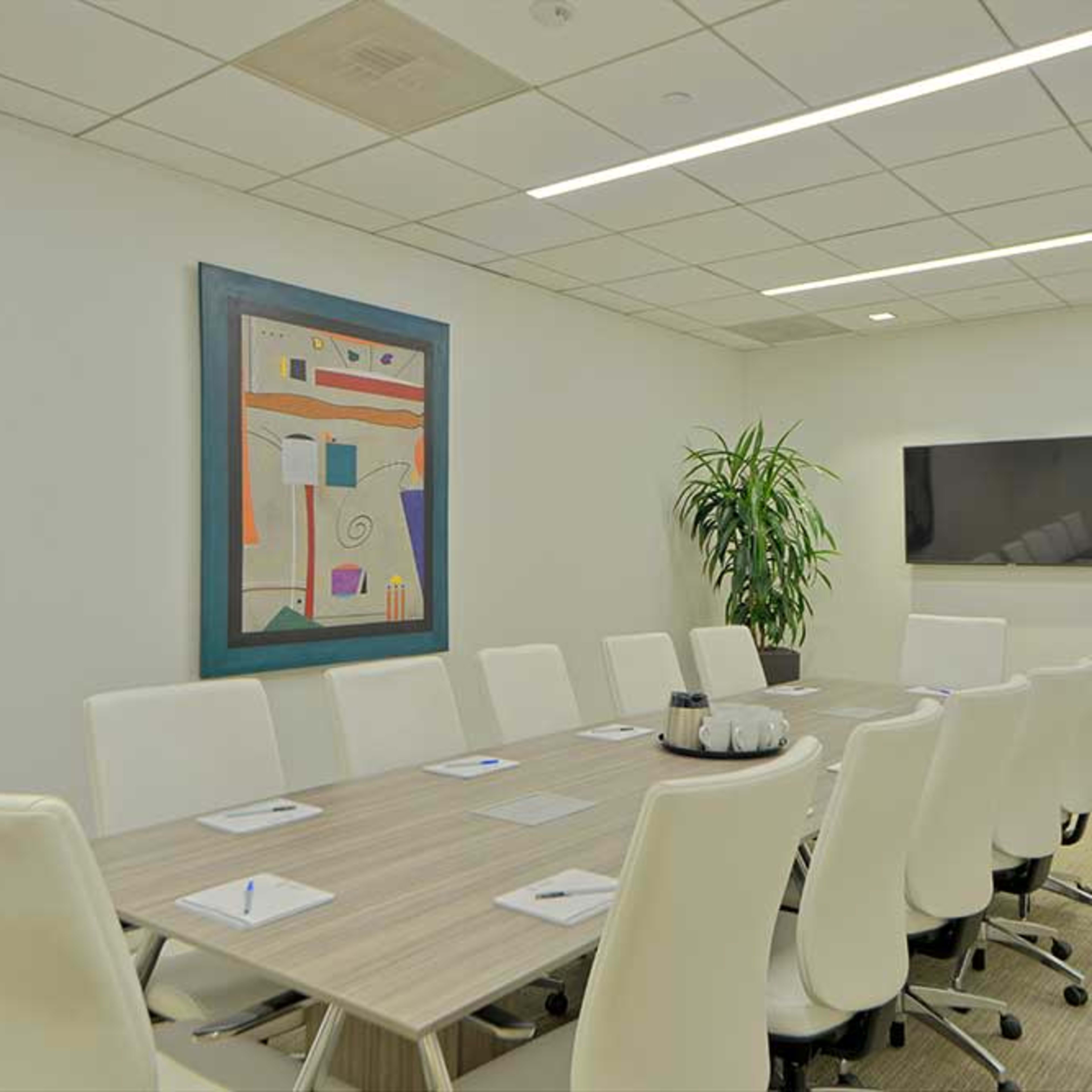 A modern conference room features a large oval table surrounded by white chairs, a wall-mounted TV, and a colorful abstract painting.