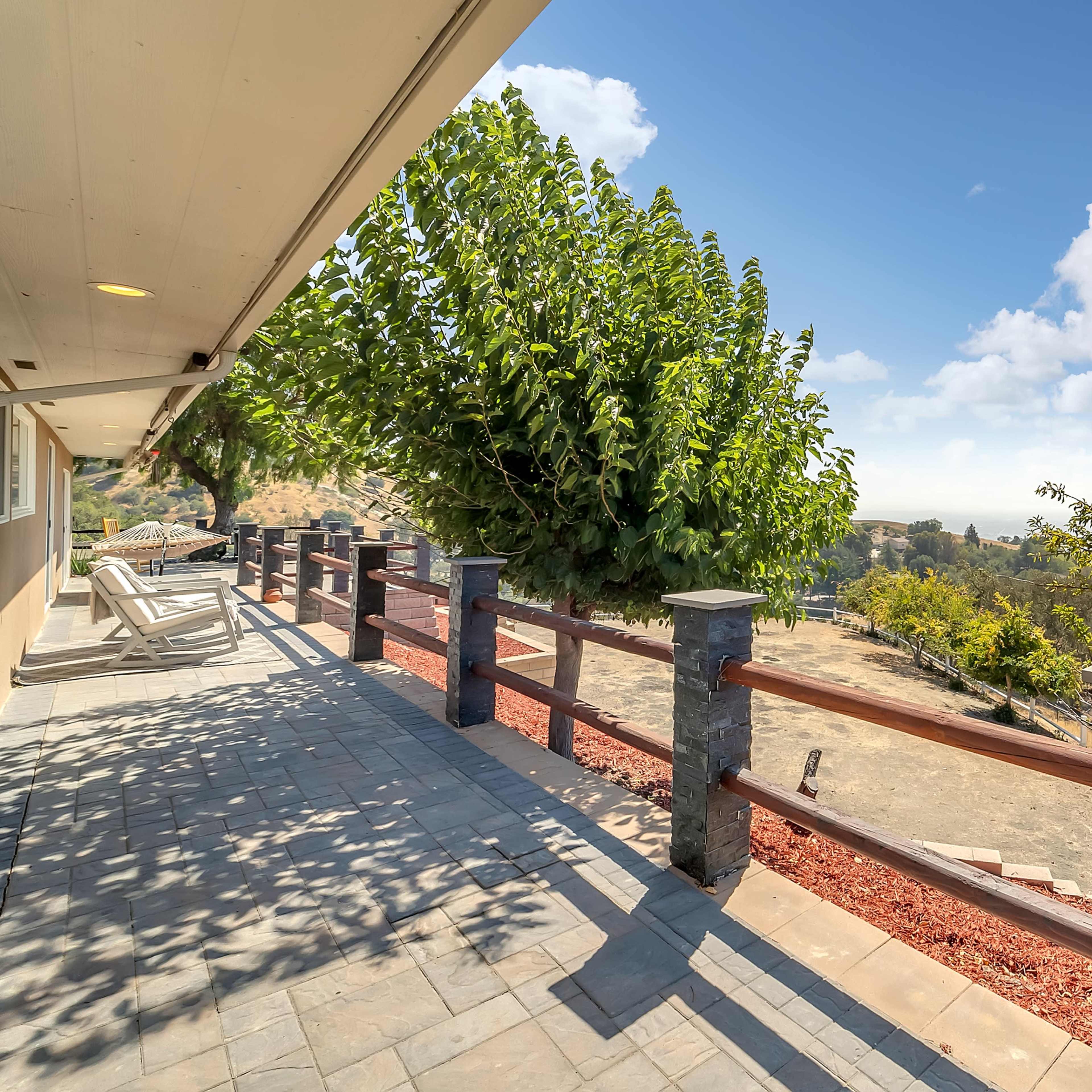 A stone patio with wooden railings overlooks a hilly landscape under a partly cloudy sky.