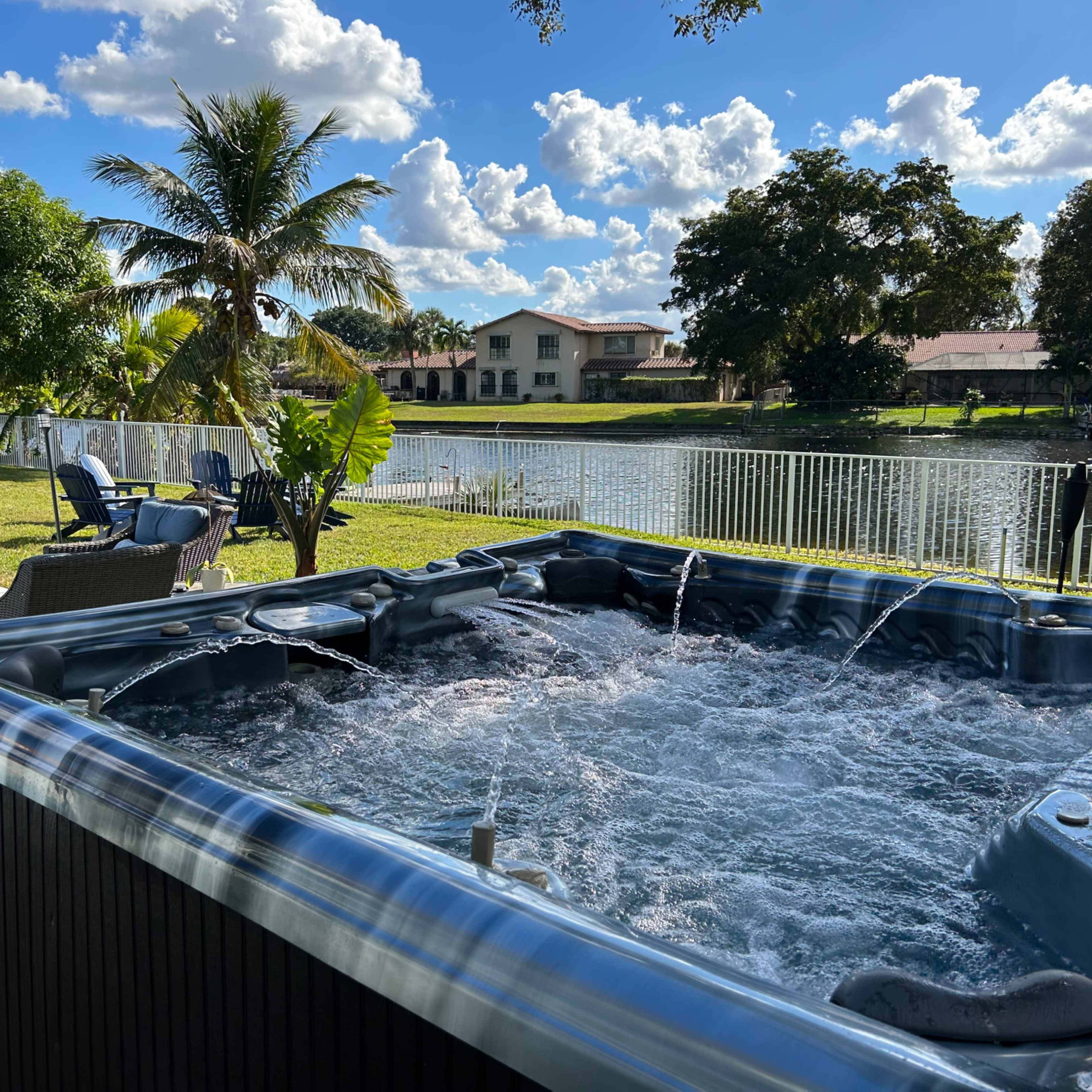 A hot tub with bubbling water is situated beside a grassy area overlooking a lake and a line of palm trees.