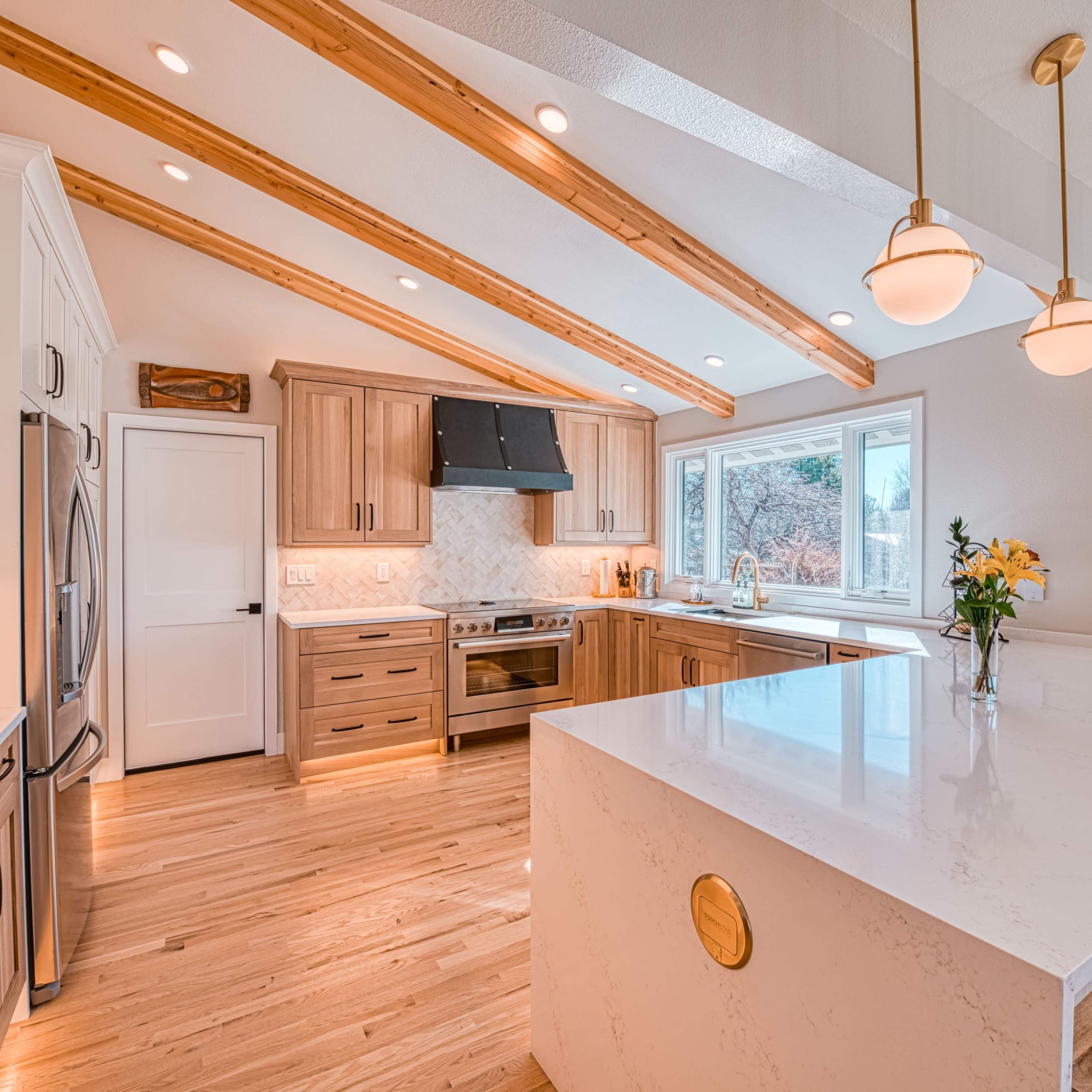 A modern kitchen features wooden beams, a large island with bar stools, and ample natural light from a window.