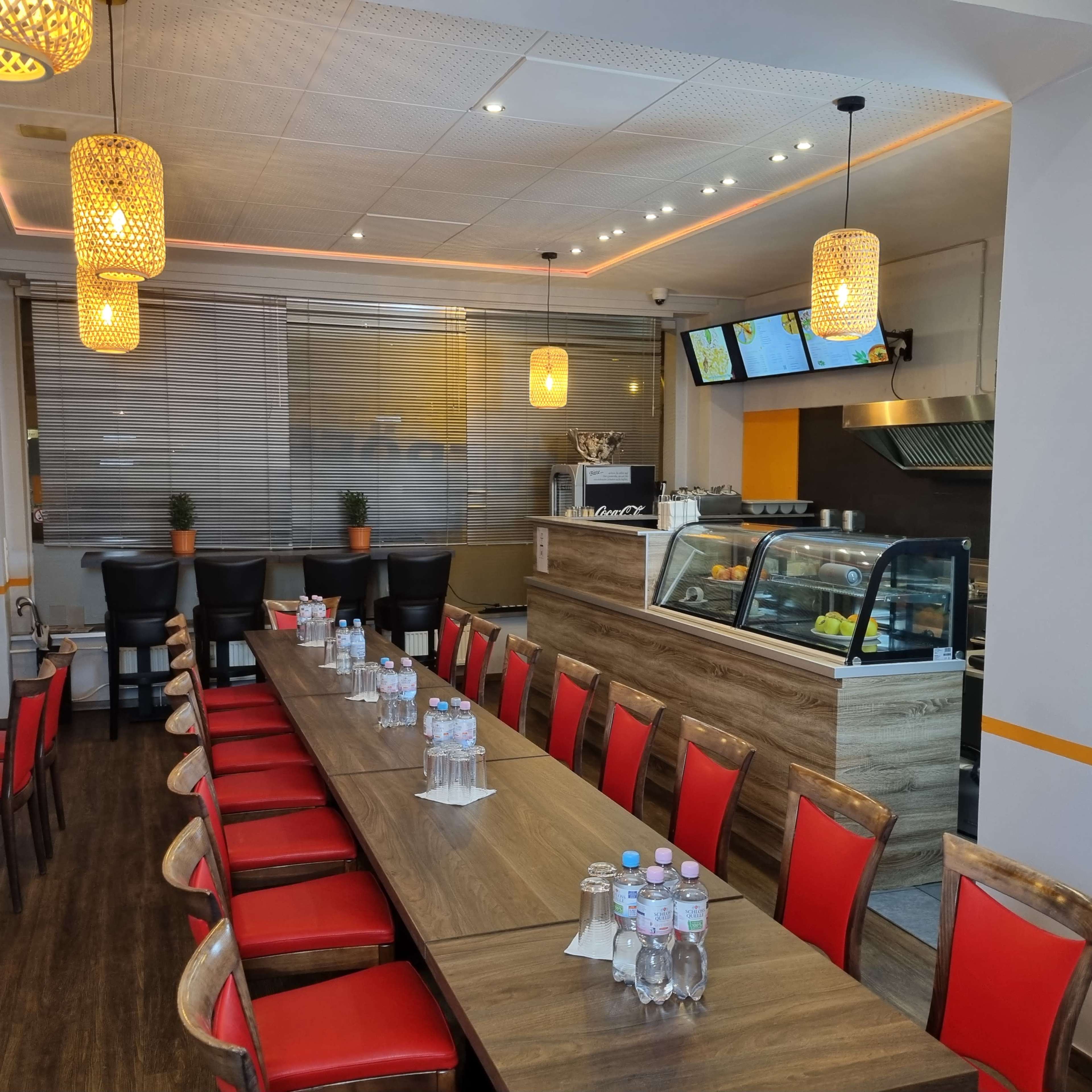 A restaurant interior with a long wooden table surrounded by red chairs, a service counter, and pendant lighting overhead.