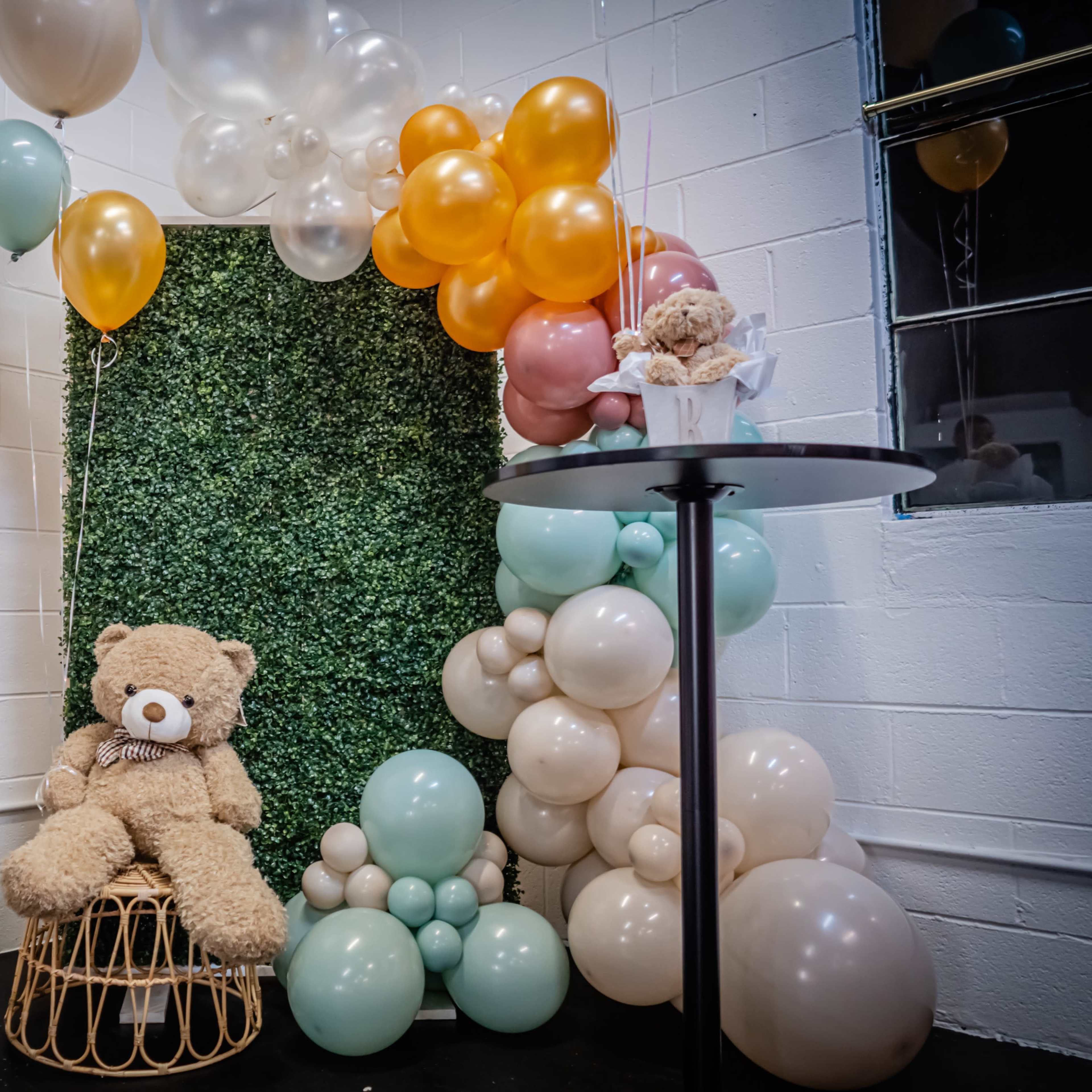 A decorated corner featuring a stuffed bear seated on a wicker chair, surrounded by colorful balloons and floral arrangements.