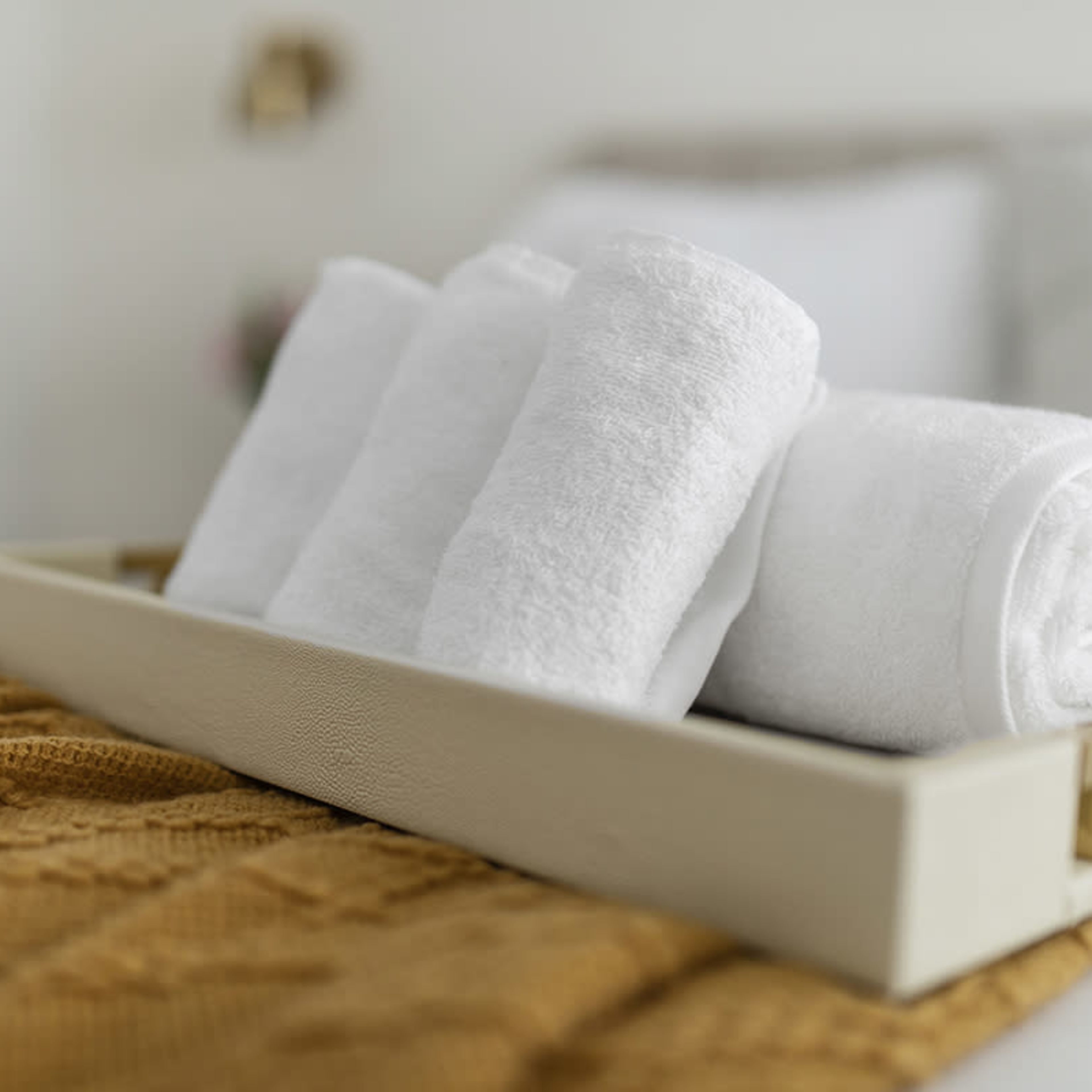 A tray holds neatly rolled white towels on a bed with a textured brown blanket.
