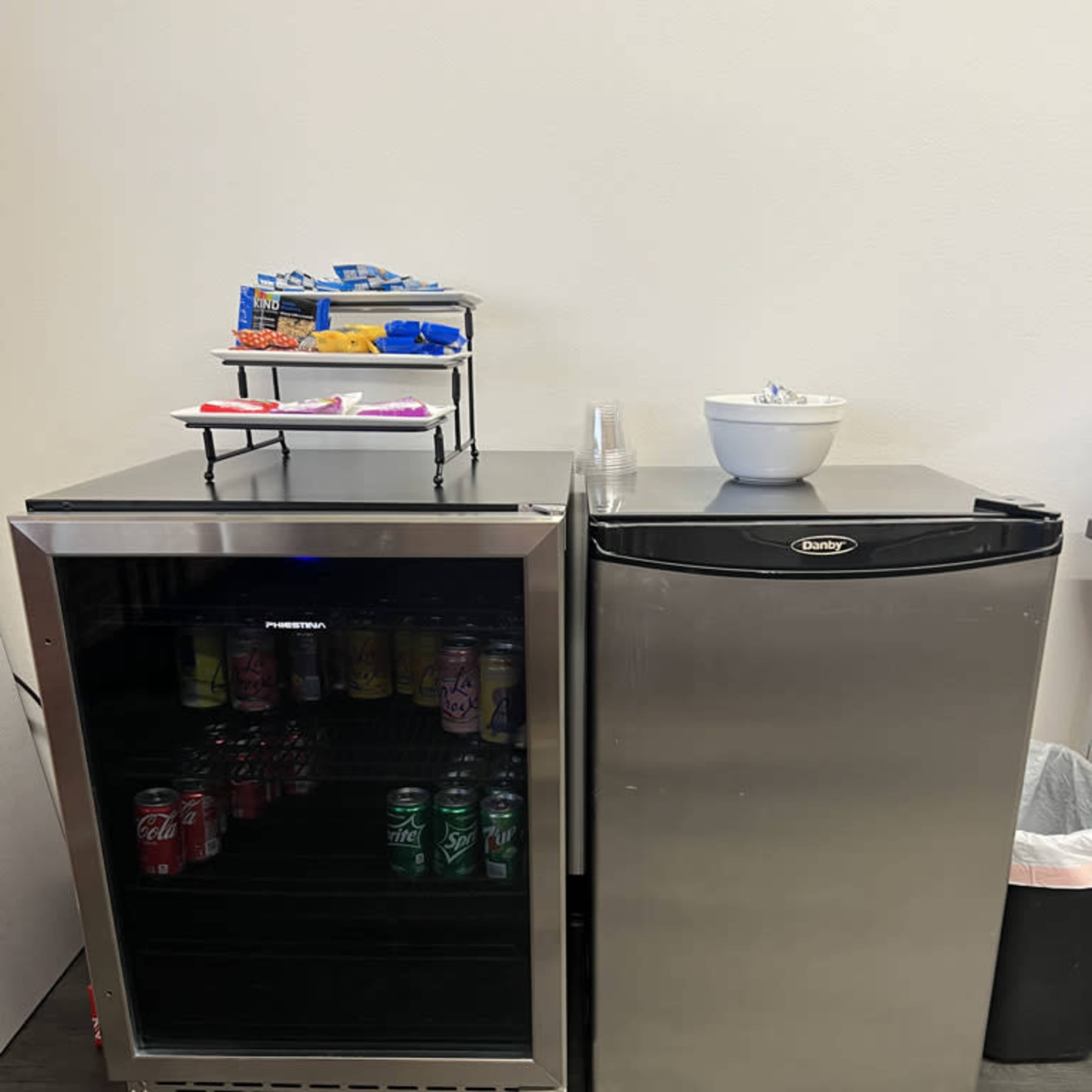 A small kitchen area featuring two compact refrigerators, one with a glass door displaying various canned drinks and the other in stainless steel, alongside a bowl and snacks on a shelf above.