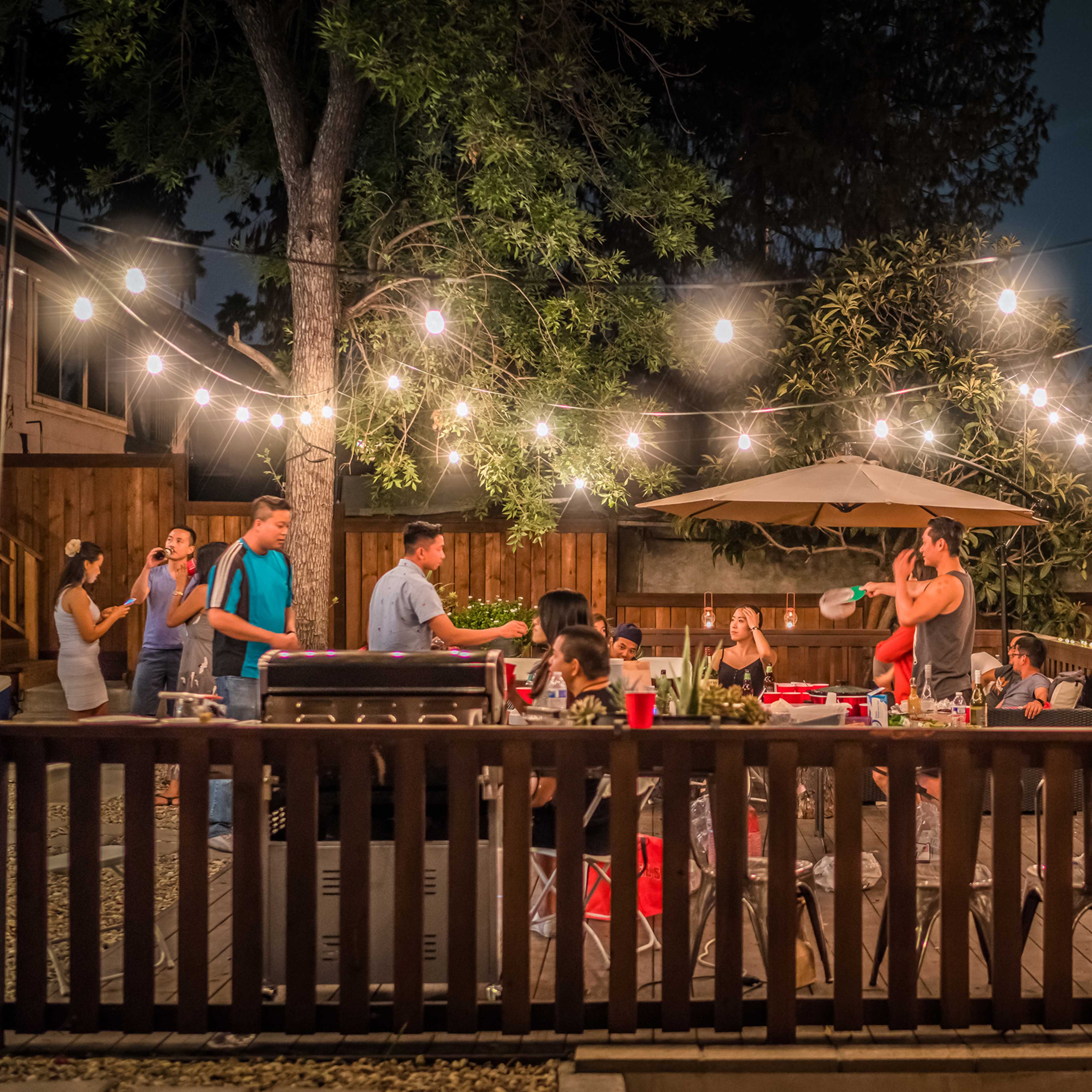 A group of people gathers around a patio with string lights in the evening, engaging in a barbecue and conversation.
