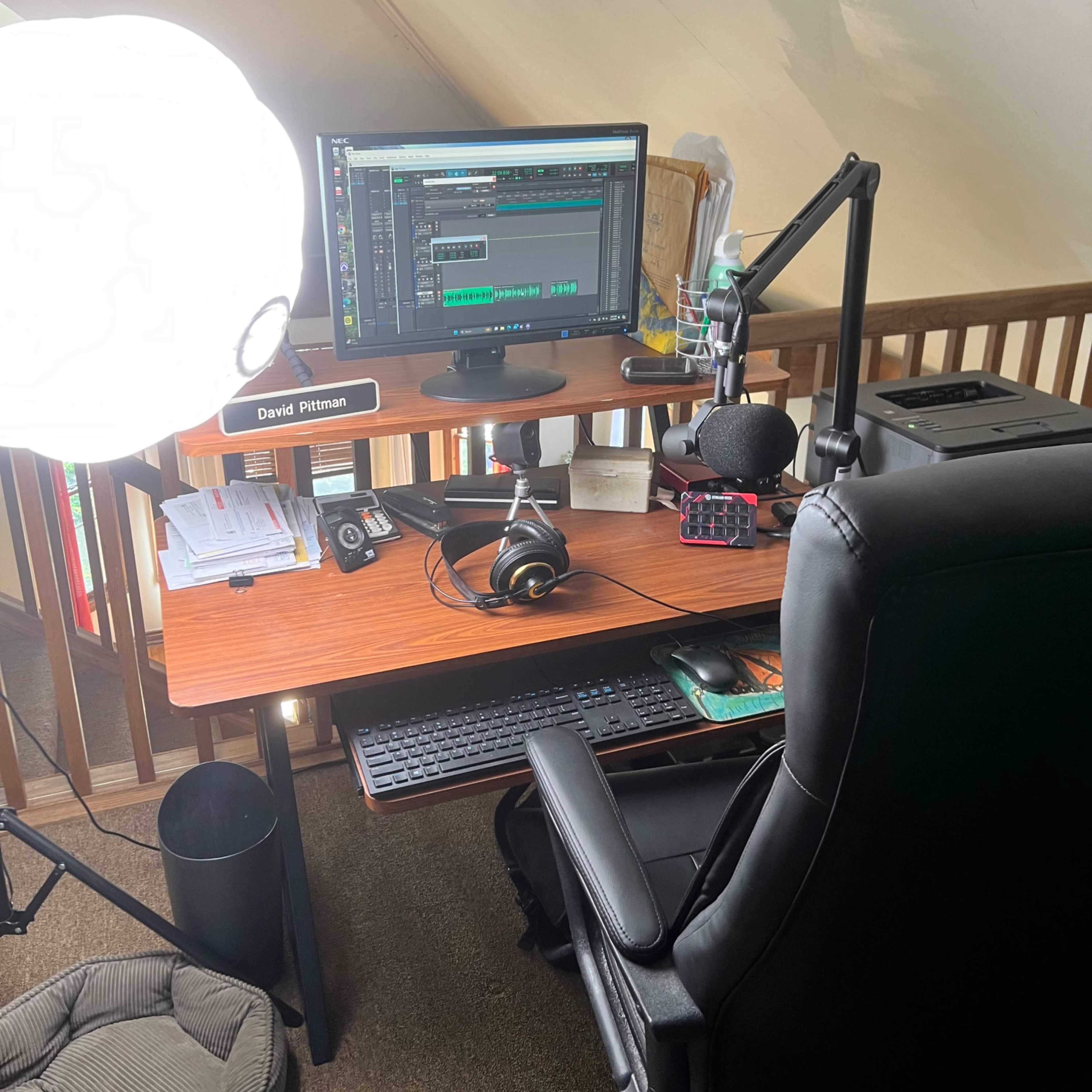 A wooden desk is set up in a loft space with a computer monitor displaying audio editing software, a microphone on a stand, headphones, and a large light source nearby.