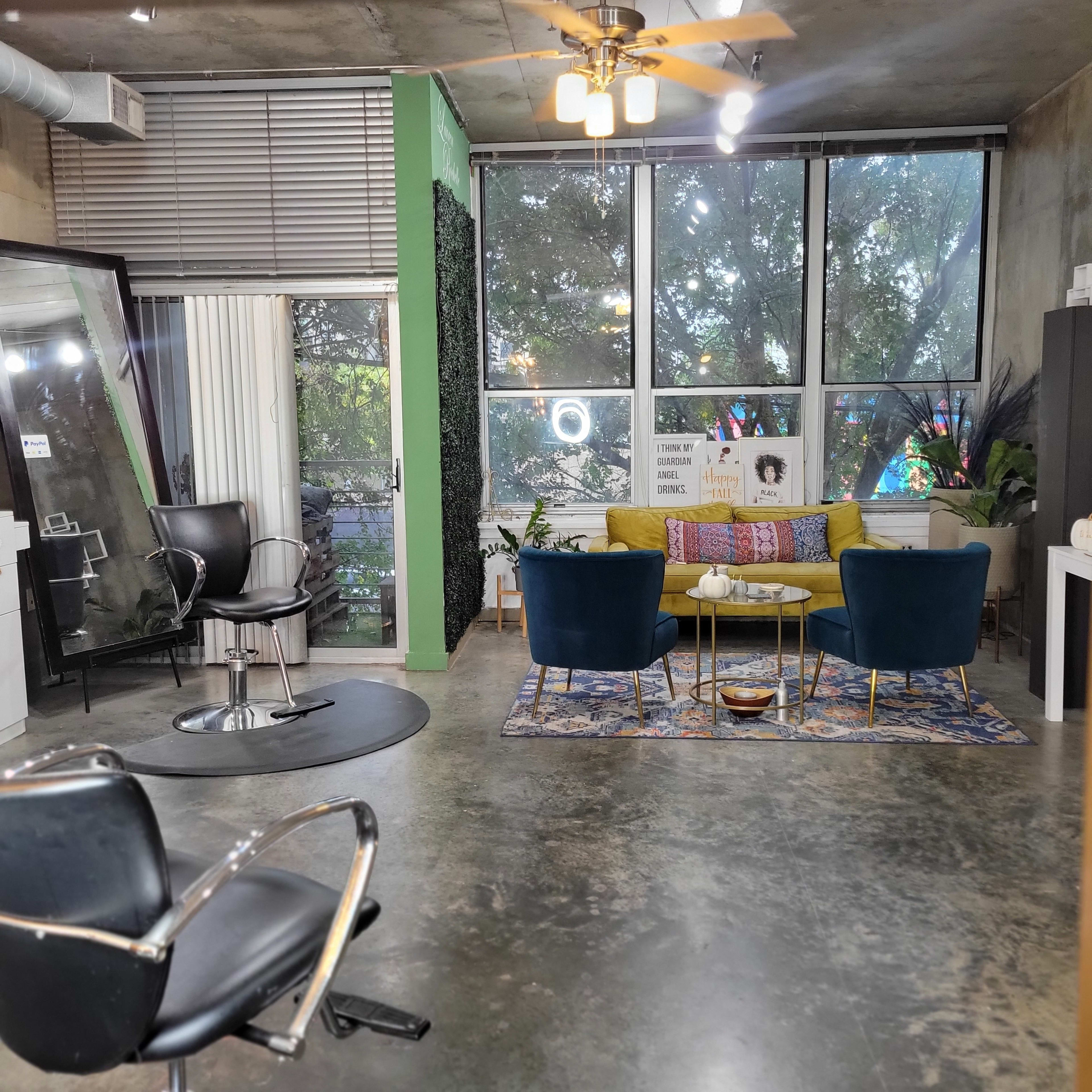 A modern salon interior with a seating area featuring a yellow couch and two blue chairs, along with a styling chair and a large mirror.