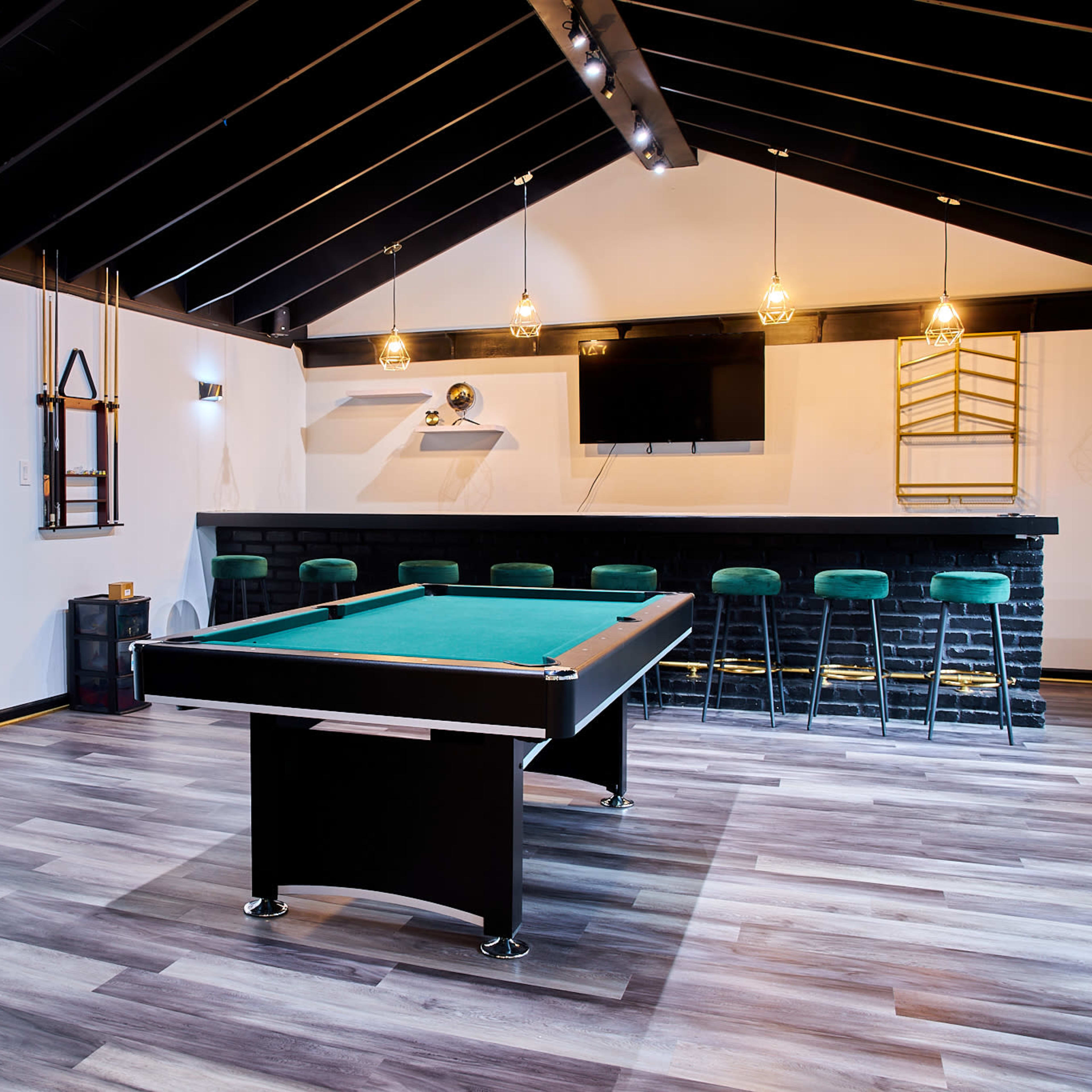 A modern game room featuring a billiards table, a bar counter with stools, and large windows allowing natural light.