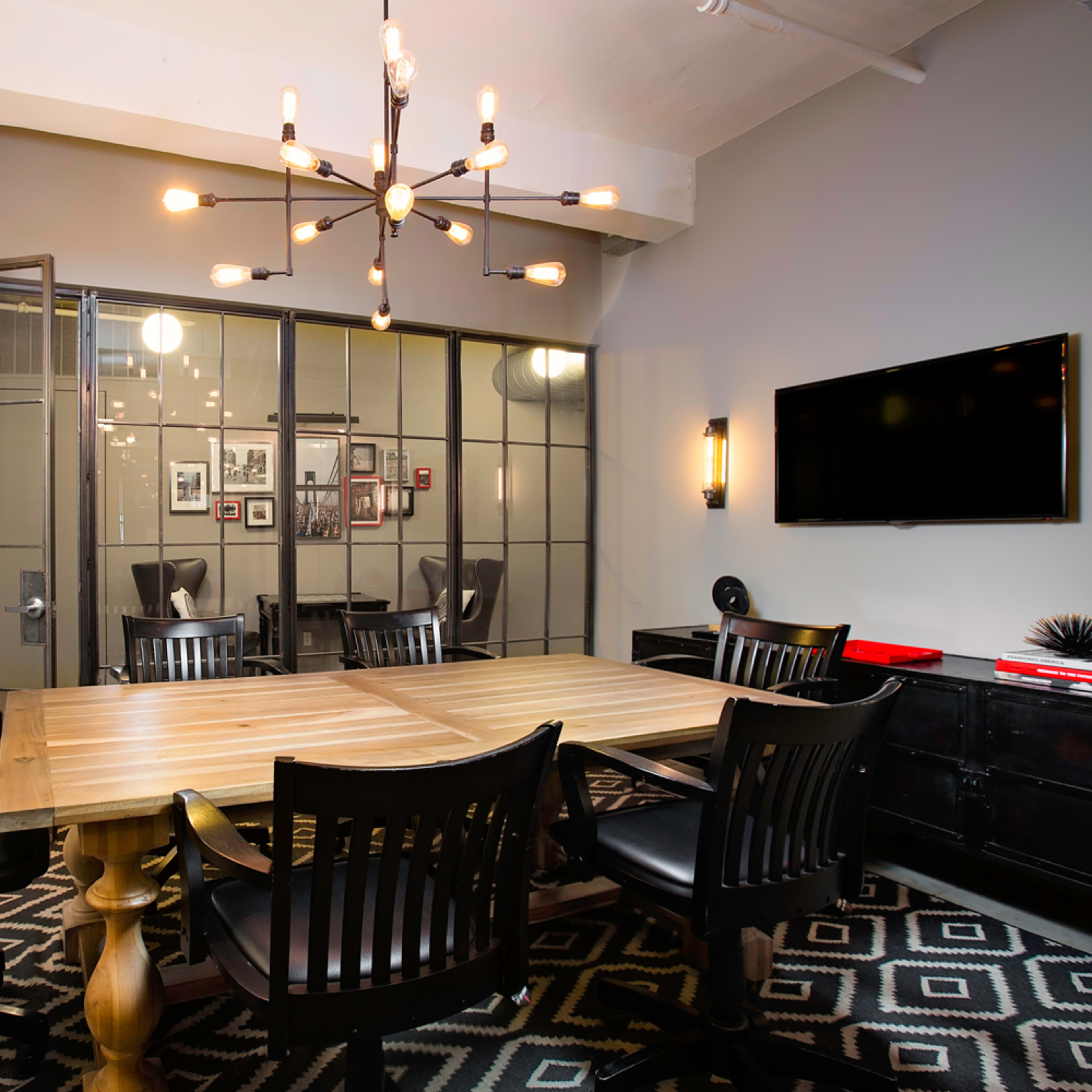 A modern conference room featuring a wooden table surrounded by black chairs, with a large glass door and wall of framed pictures in the background.