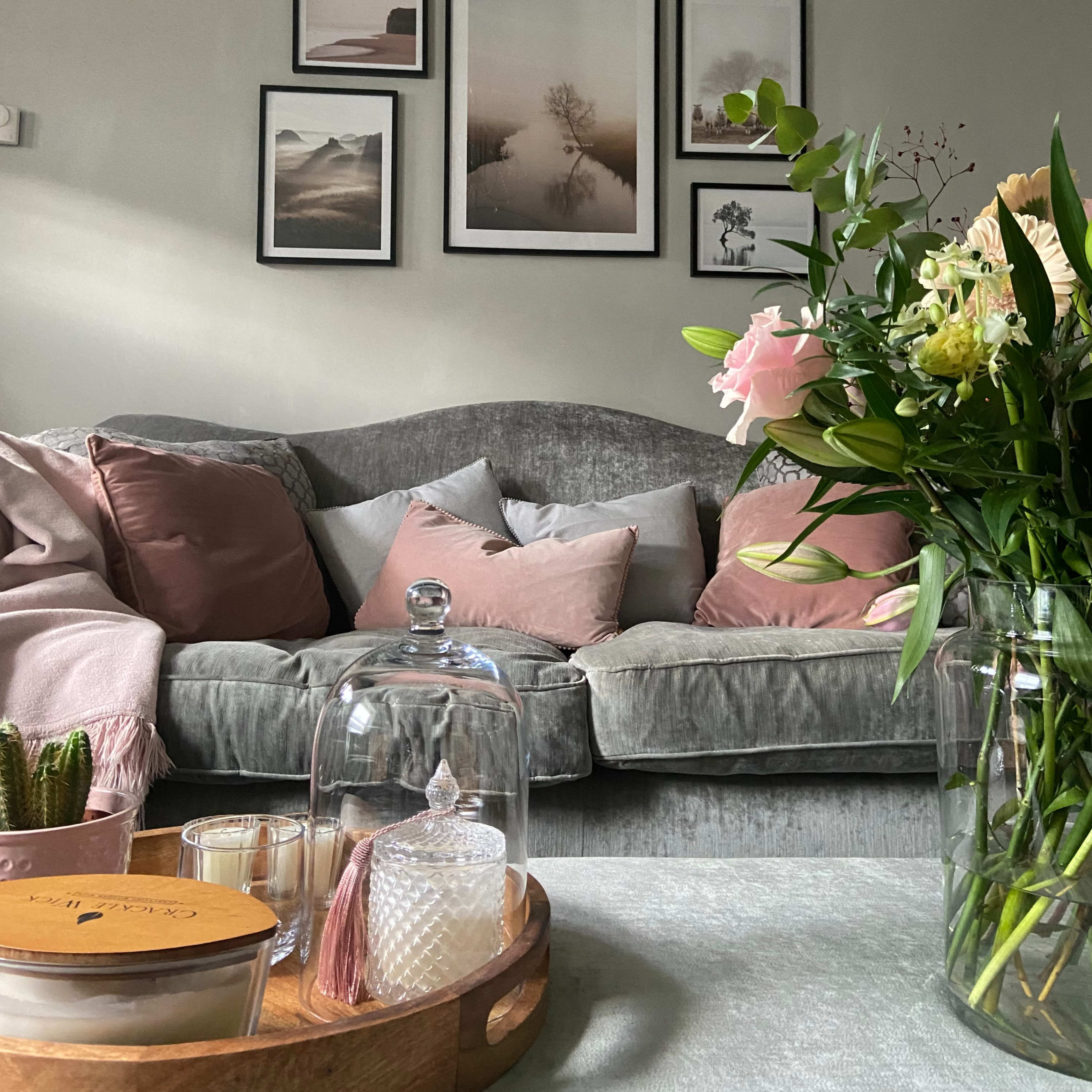 A cozy living room features a gray sofa adorned with pink cushions, framed photographs on the wall, and a coffee table with decorative items and a vase of flowers.