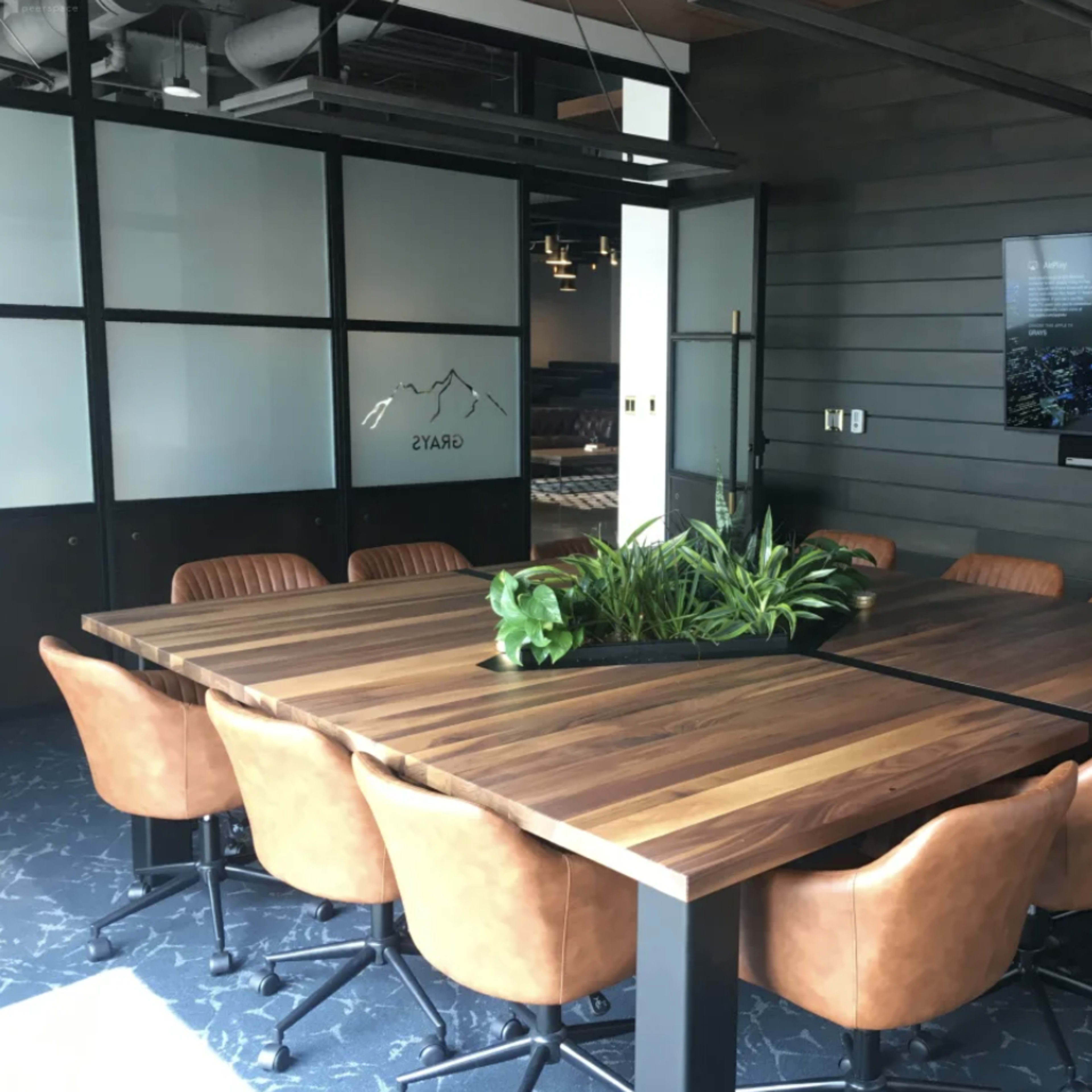 A large wooden conference table surrounded by brown leather chairs is positioned in a modern meeting room with a television mounted on the wall.