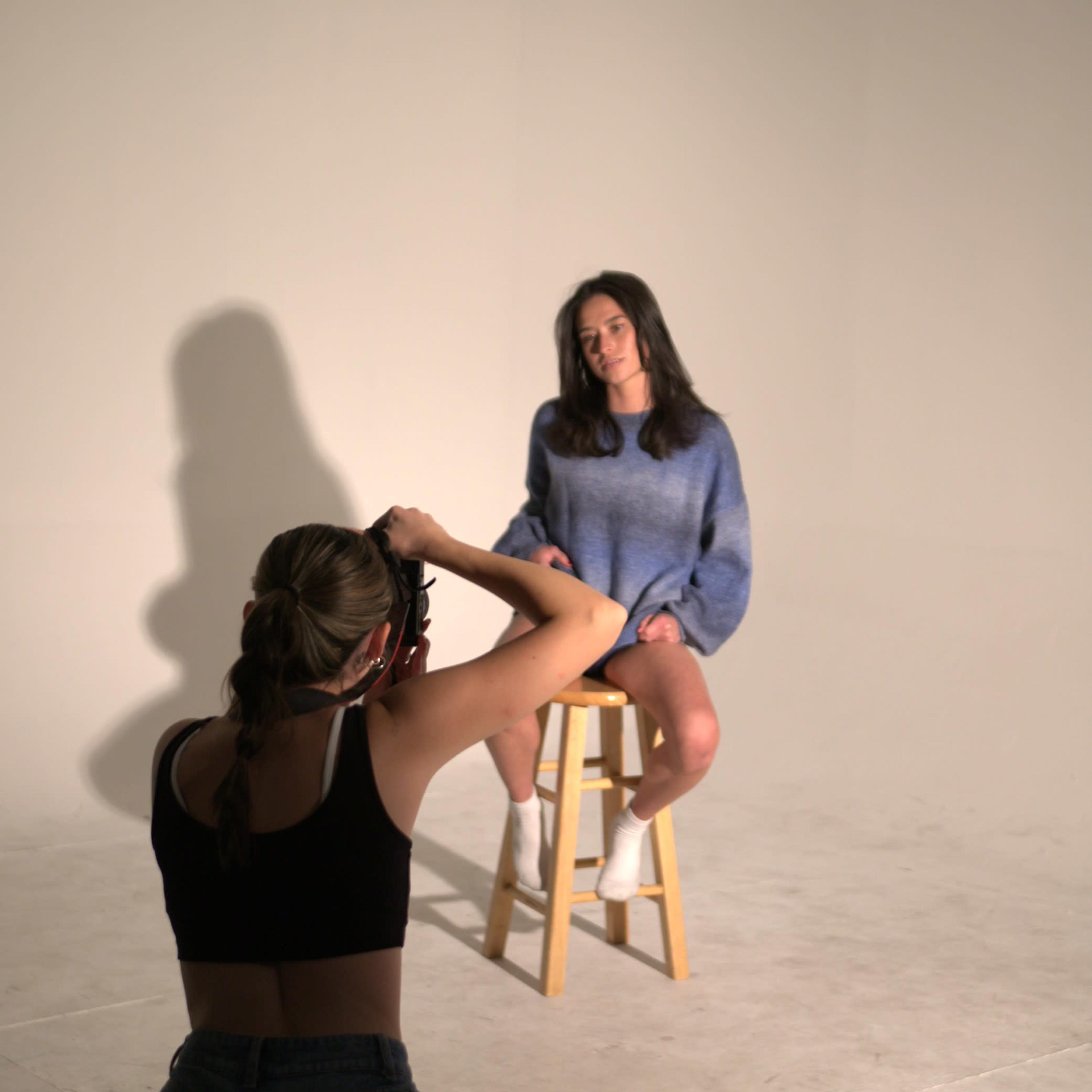 A person is taking a photograph of a seated model on a stool in a plain studio setting.