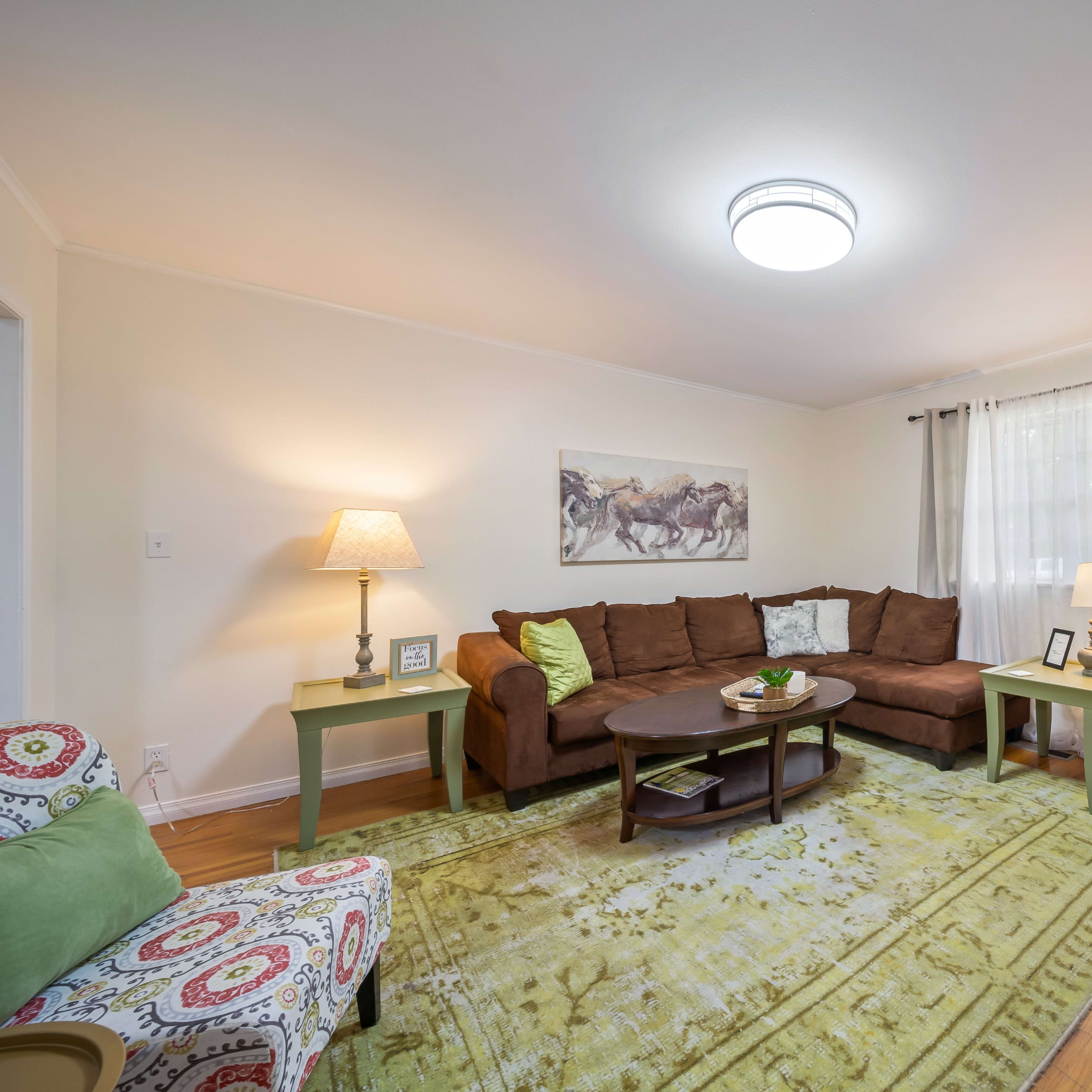 A cozy living room featuring brown sectional furniture, light green accents, a round coffee table, and a large area rug on wooden flooring, with natural light coming through sheer curtains.