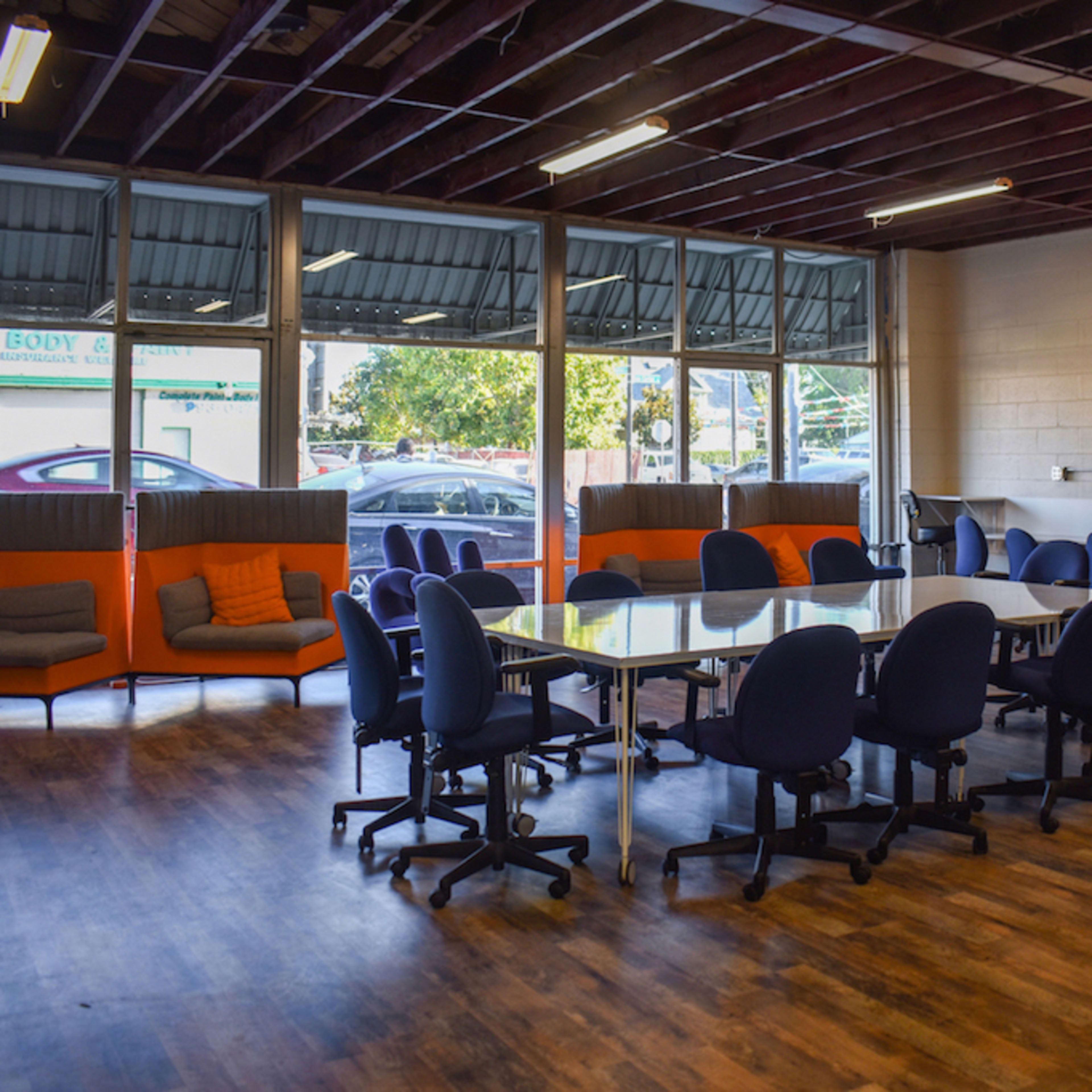 A large room features a conference table surrounded by rolling chairs, with orange seating areas and a kitchen space visible in the background.