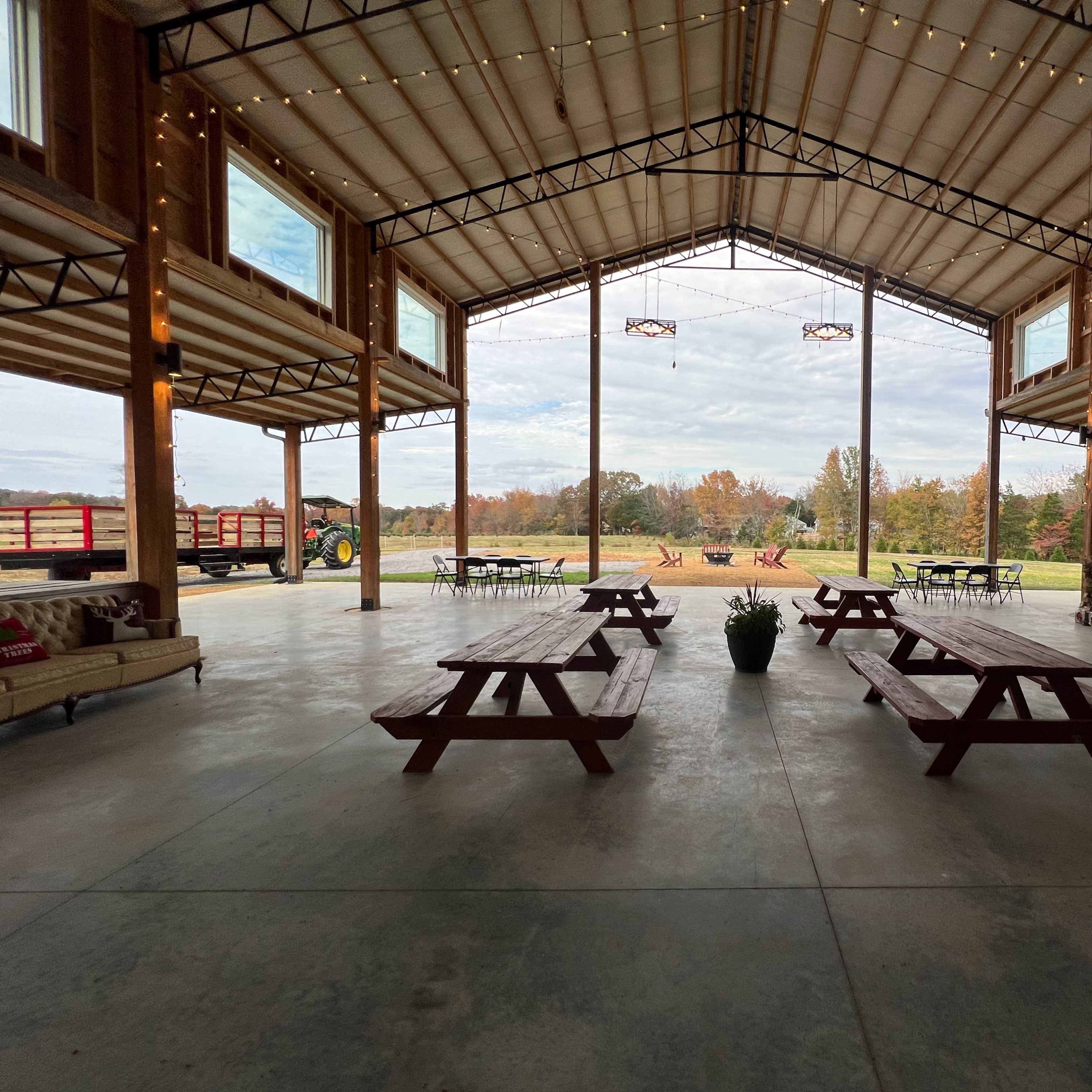 A spacious barn interior with wooden picnic tables, large windows, and a view of a landscaped outdoor area with a tractor and firewood stacks.