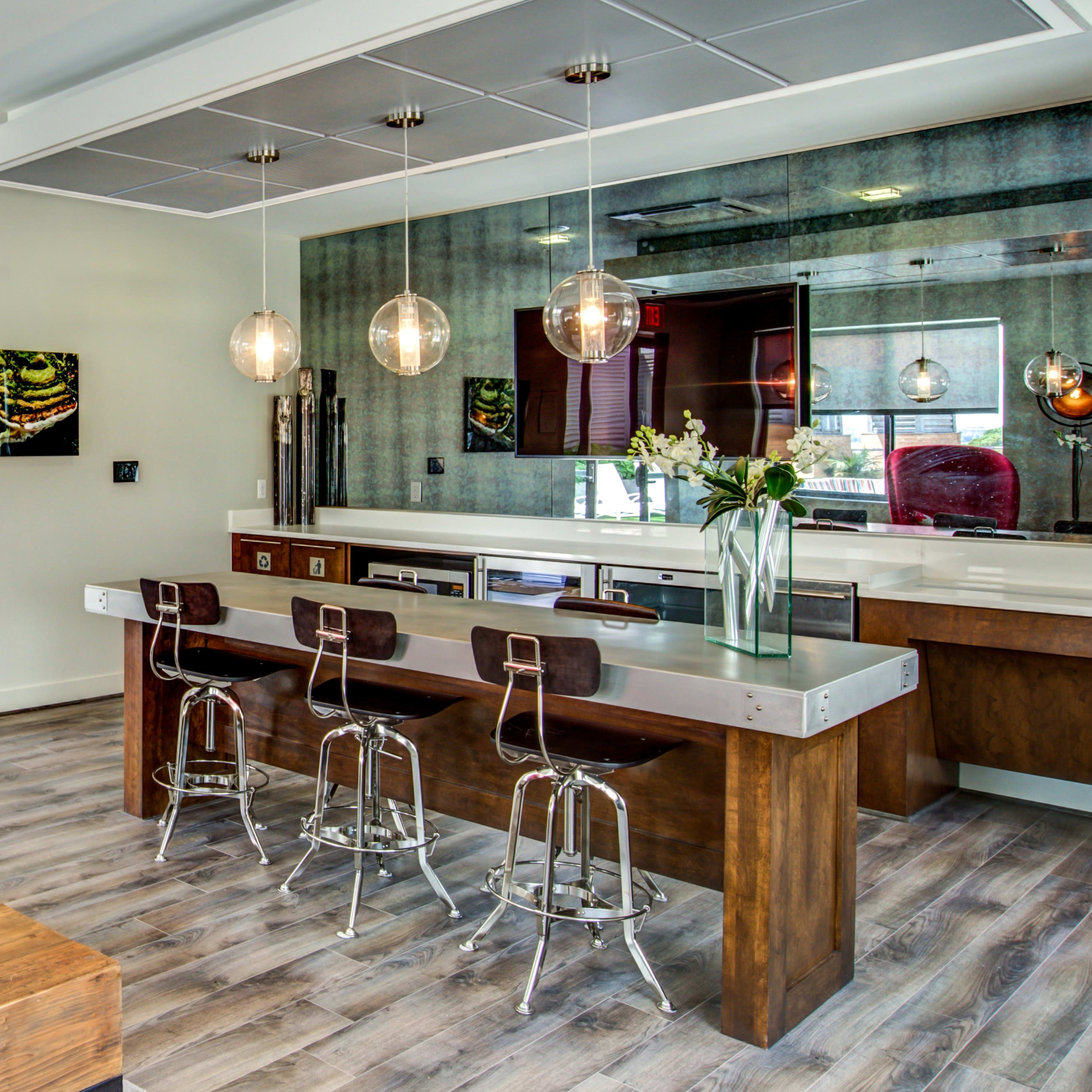 A modern kitchen and dining area featuring a long countertop with bar stools, a large mirror, and a wooden table with decorative plants.