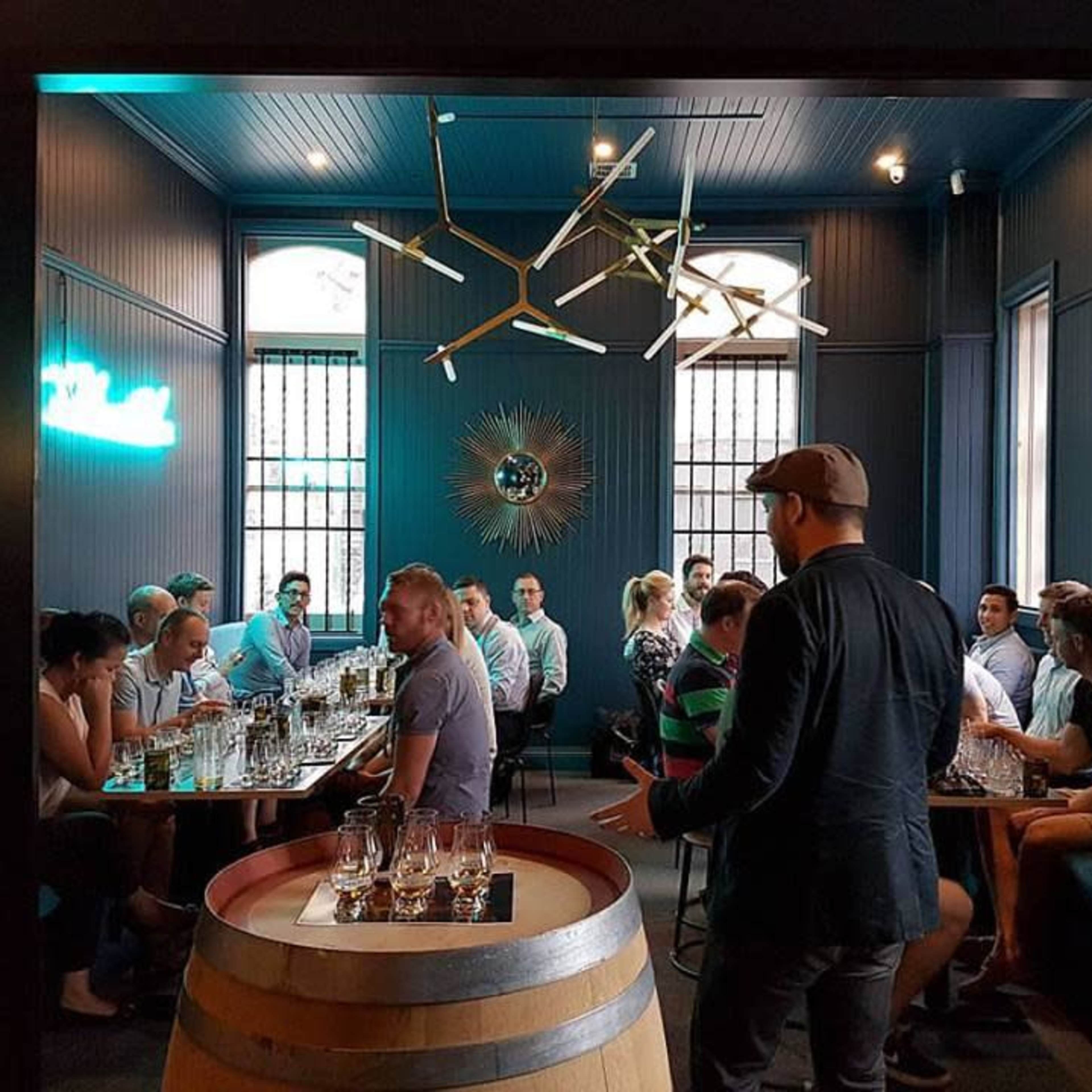 A group of people sits around tables in a dimly lit room with large windows and stylish decor, while one man stands near a barrel in the foreground.