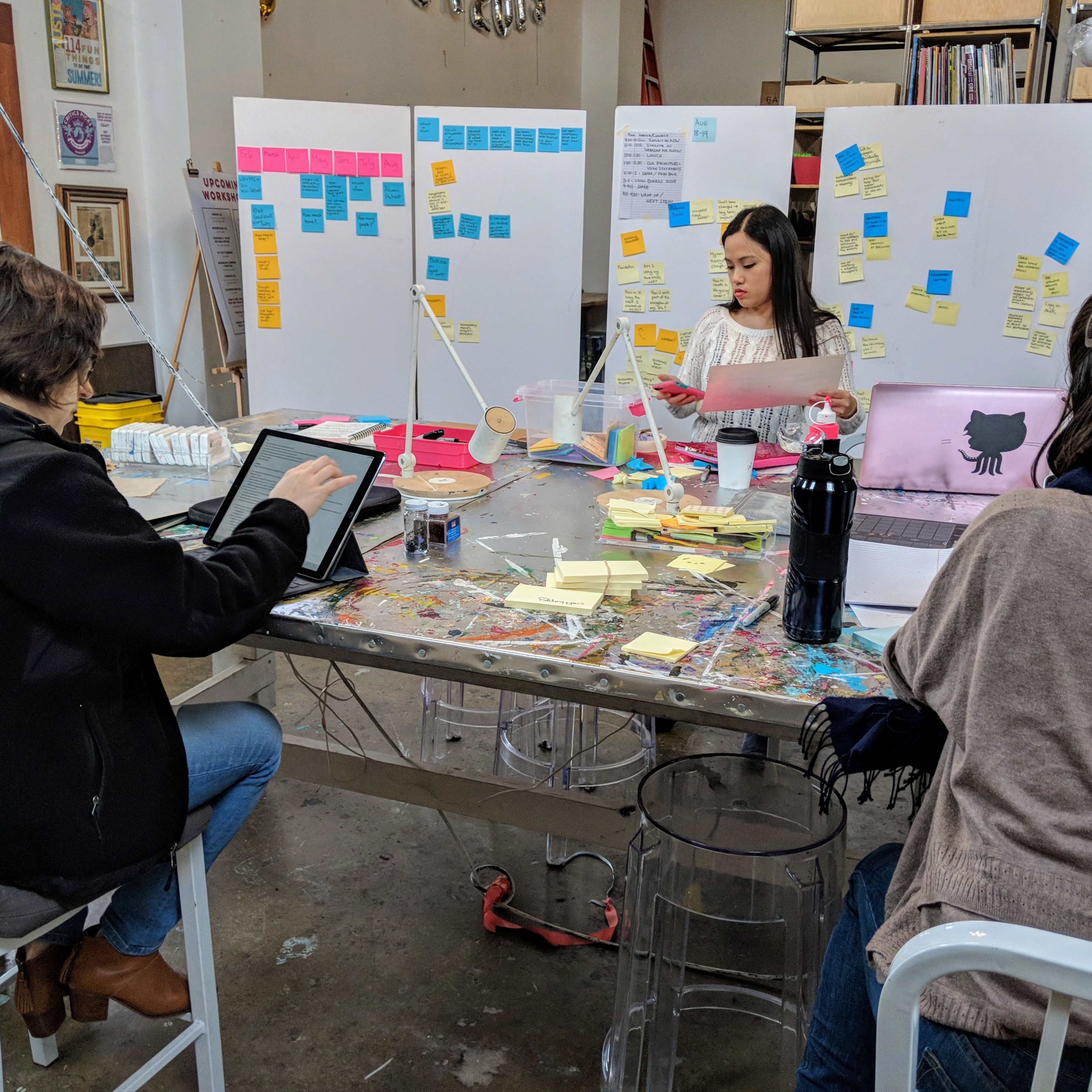 Three people collaborate in a studio filled with sticky notes on boards and a cluttered table covered in various materials.