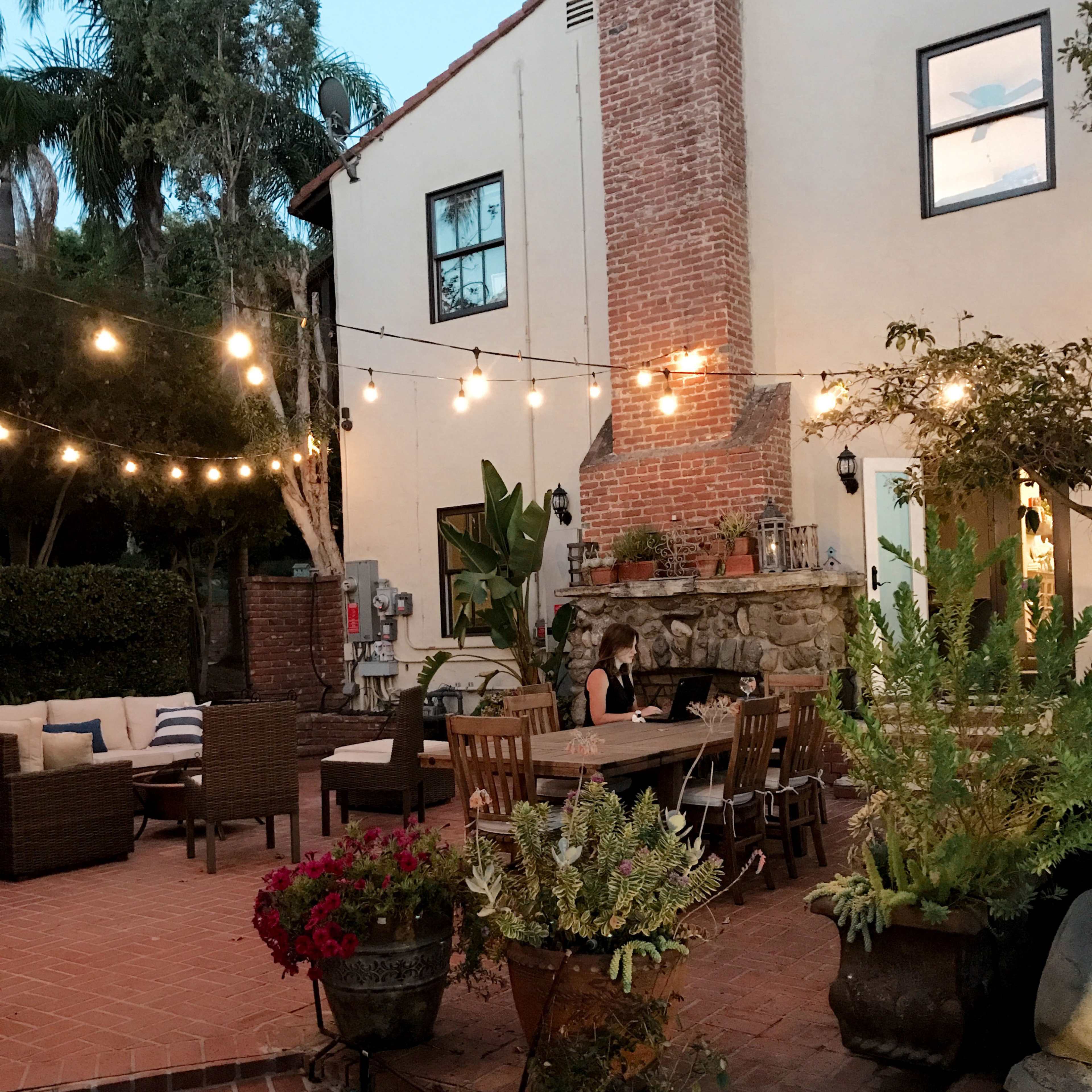 A patio area features seating around a central table, with string lights illuminating the space and greenery in decorative pots.