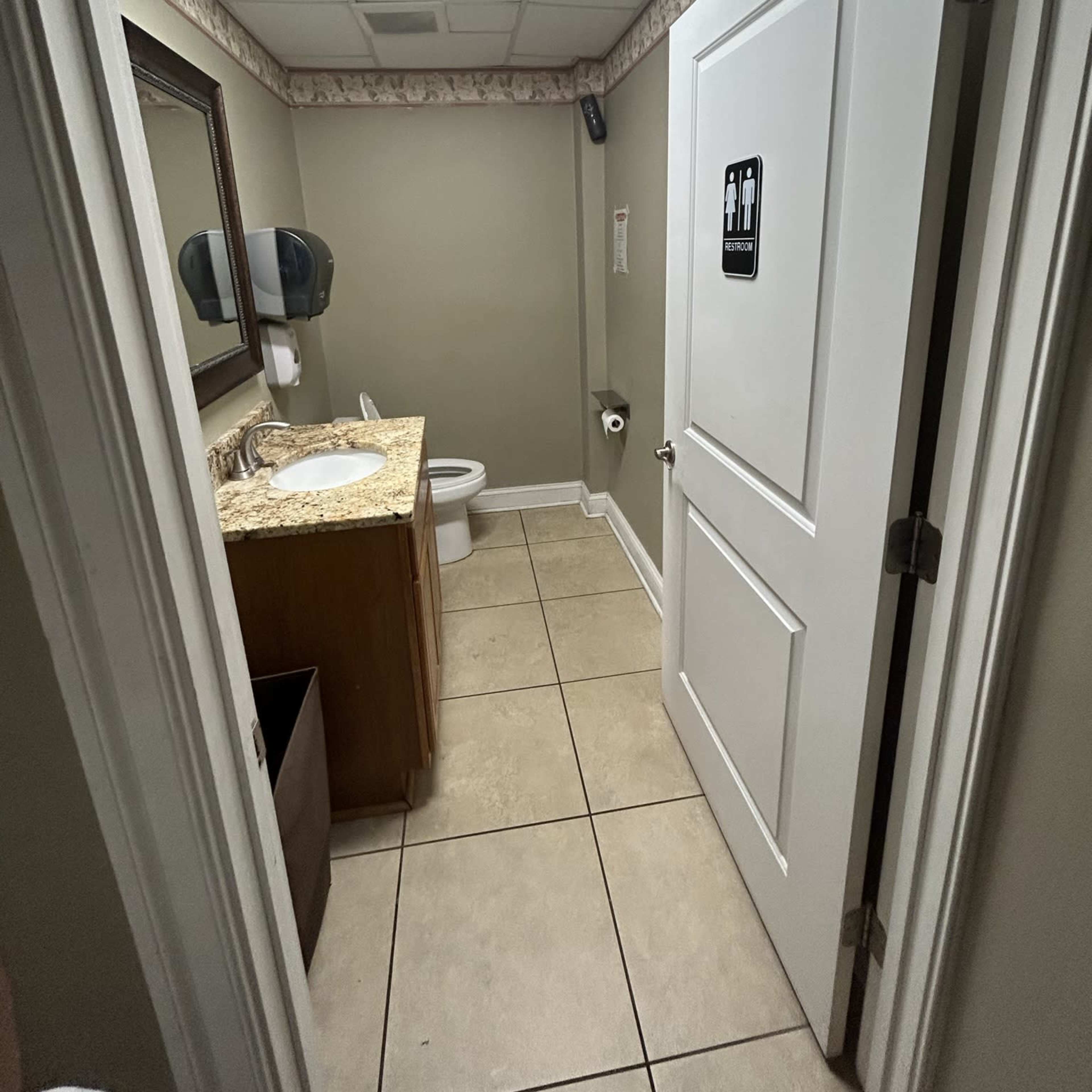 A small bathroom with a sink, mirror, toilet, and a door leading out, all within a tiled space.