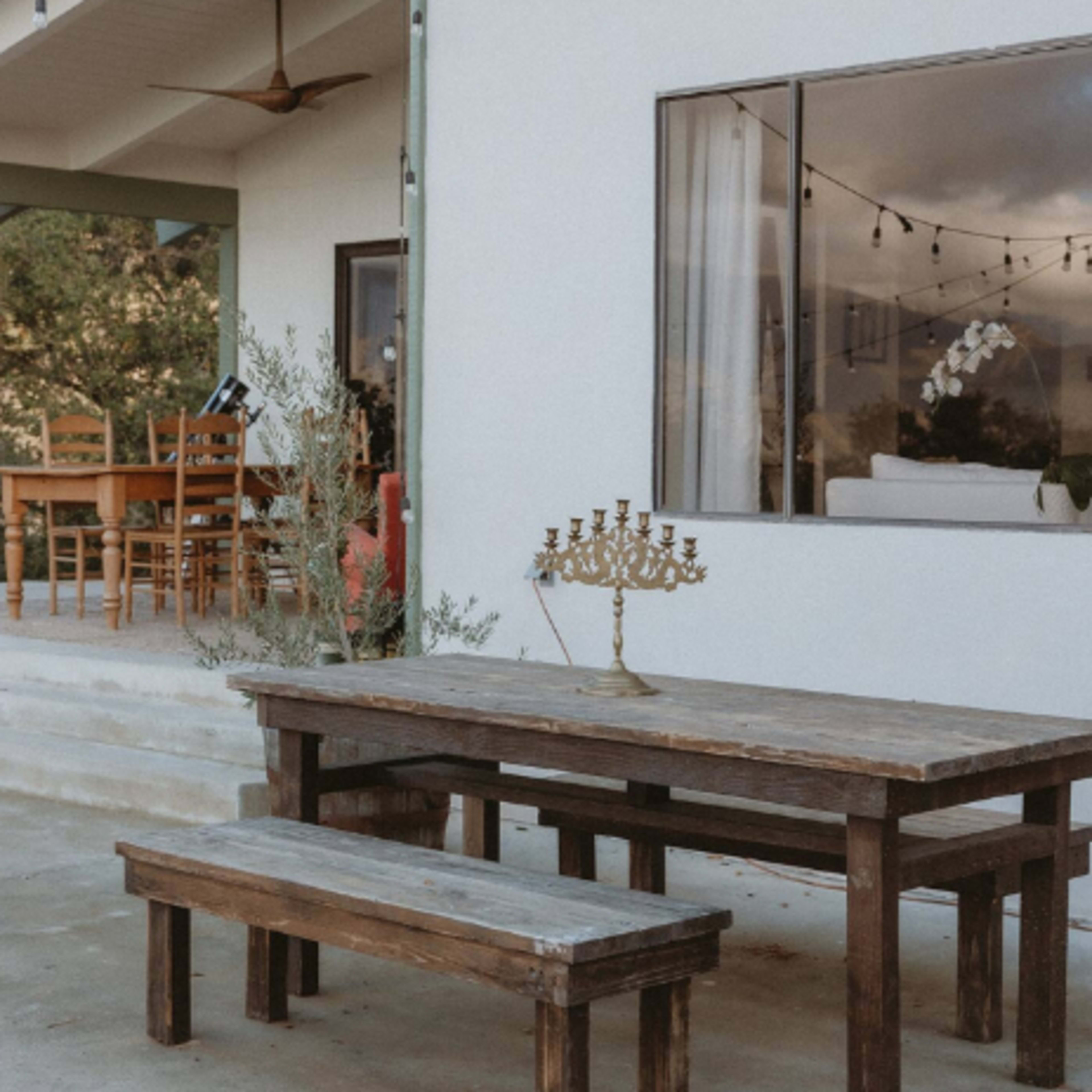 A wooden table with a matching bench and a menorah is situated outside a house with large windows and string lights overhead.