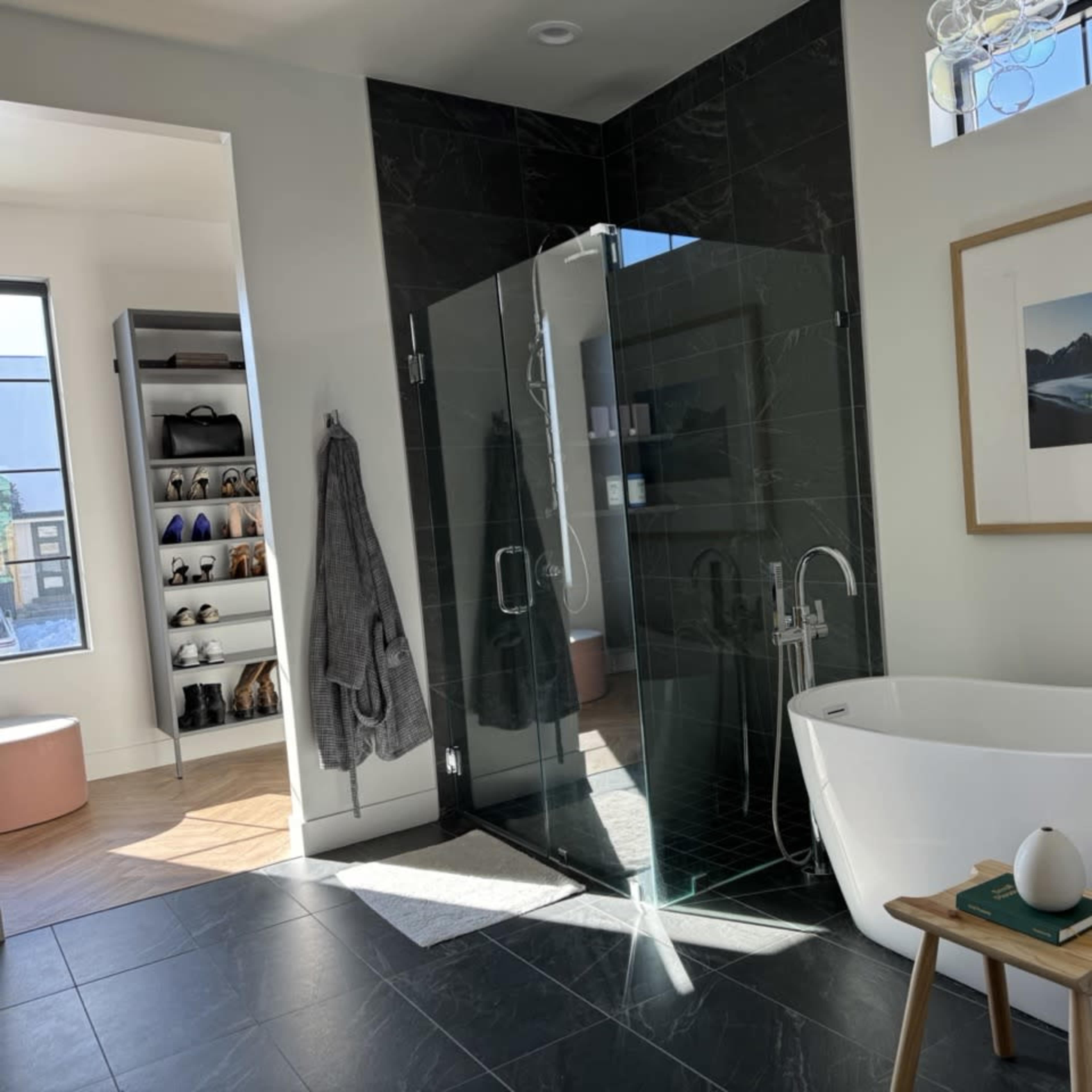 A modern bathroom featuring a glass-enclosed shower, a freestanding bathtub, a wooden stool with a decorative item, and a large window providing natural light.