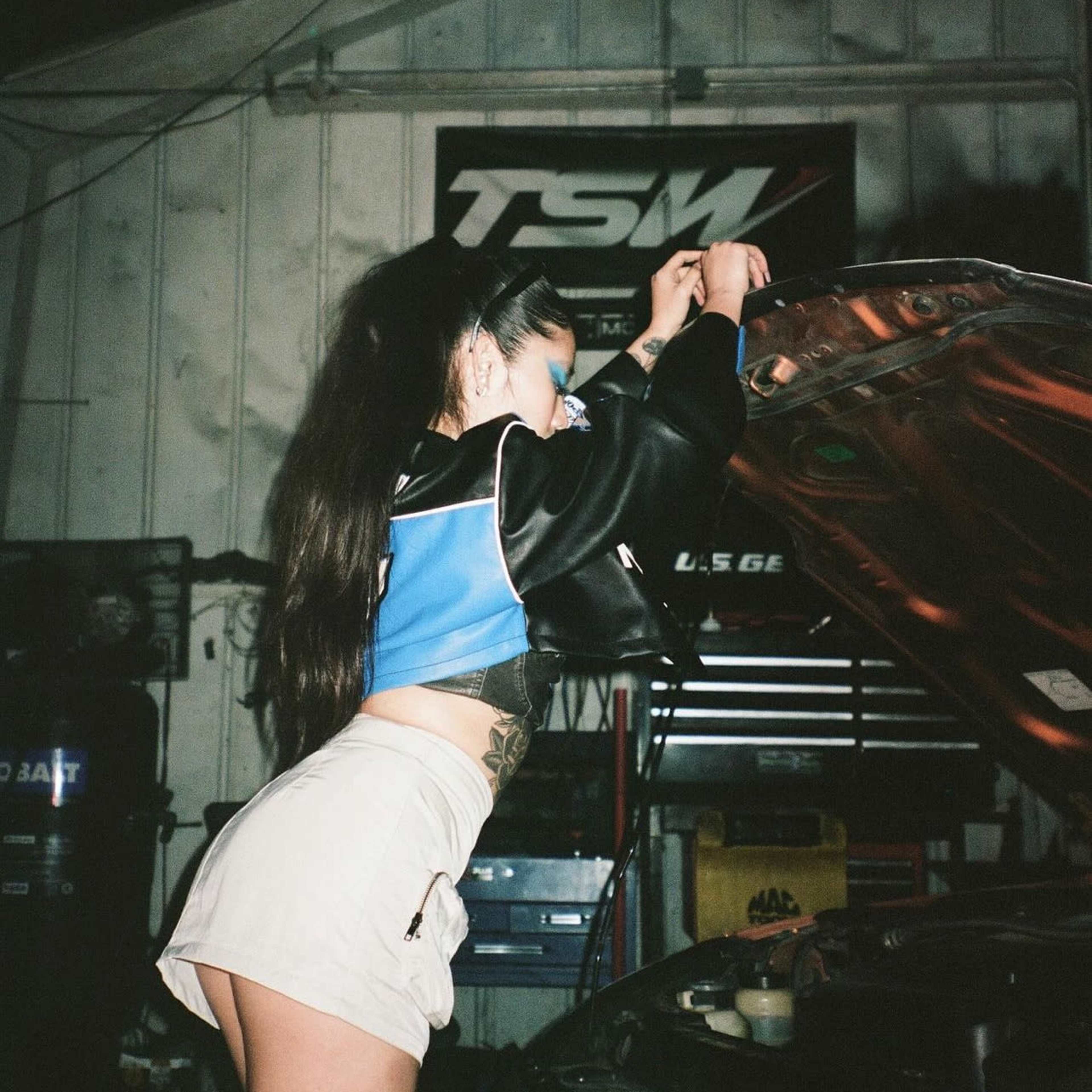 A person is leaning over the open hood of a car in a garage, working on the engine.