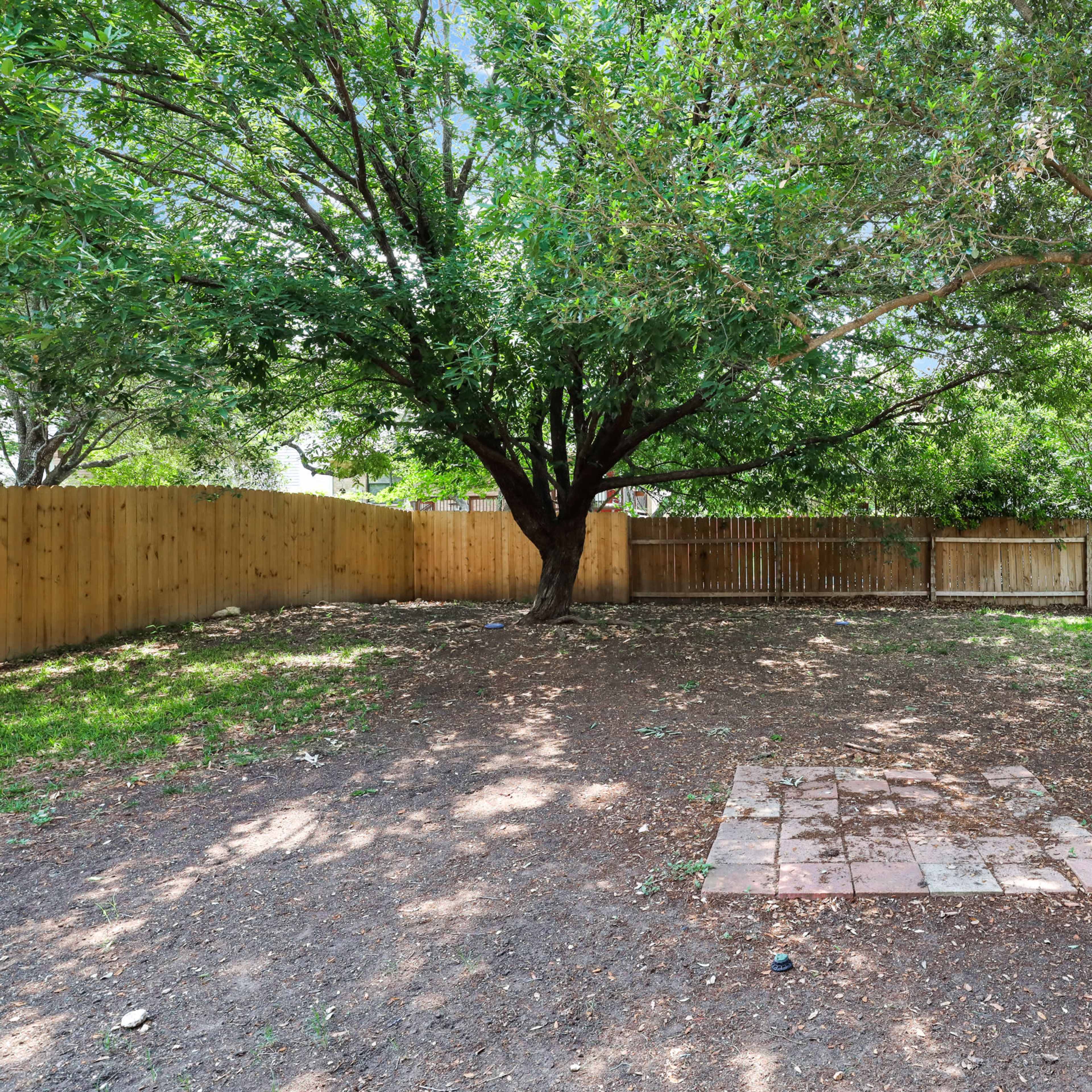 A spacious backyard features two large trees and a stone patio area surrounded by a wooden fence.