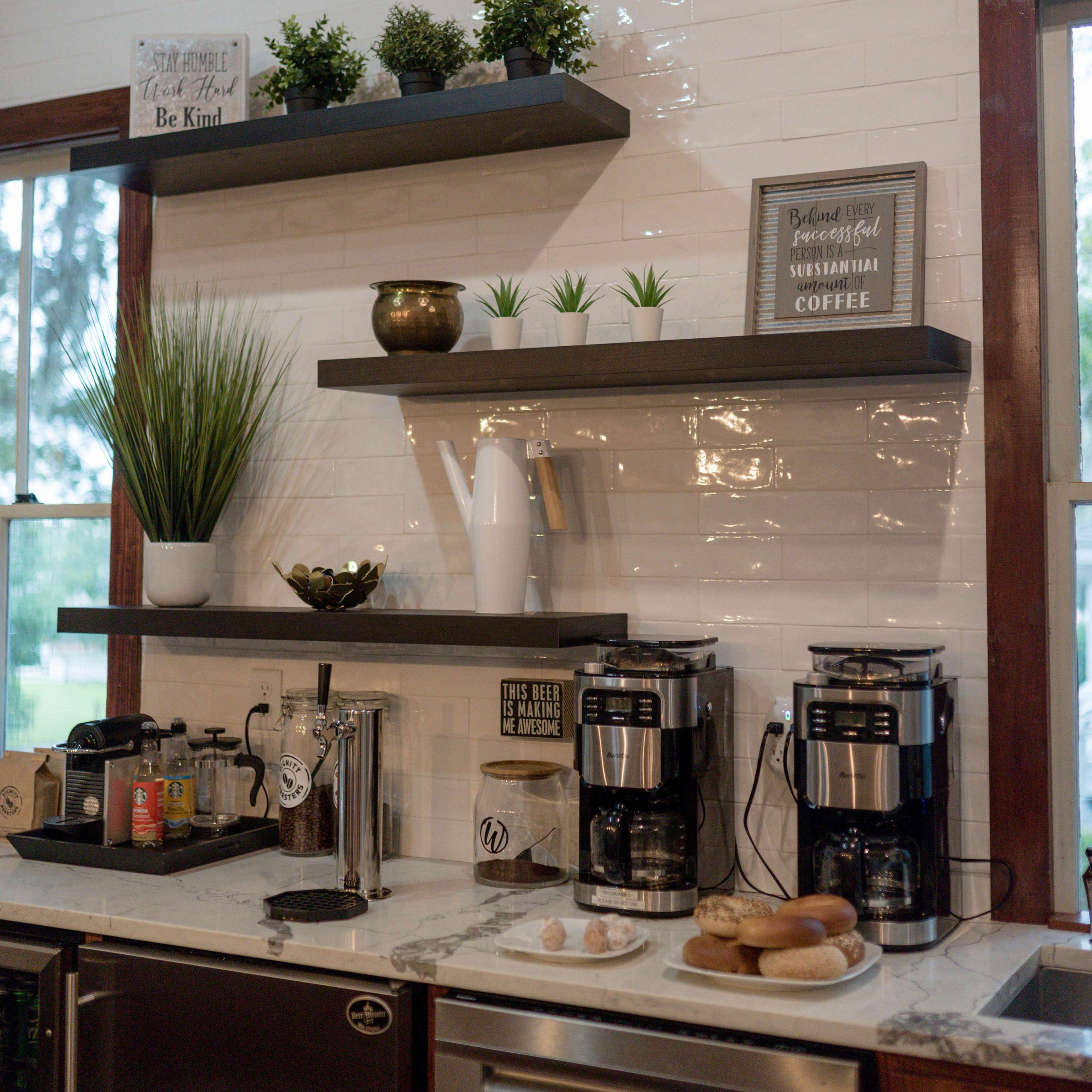 A modern kitchen with a white tiled backsplash, displaying shelves with plants and decorative items above two coffee machines and a stainless steel refrigerator.