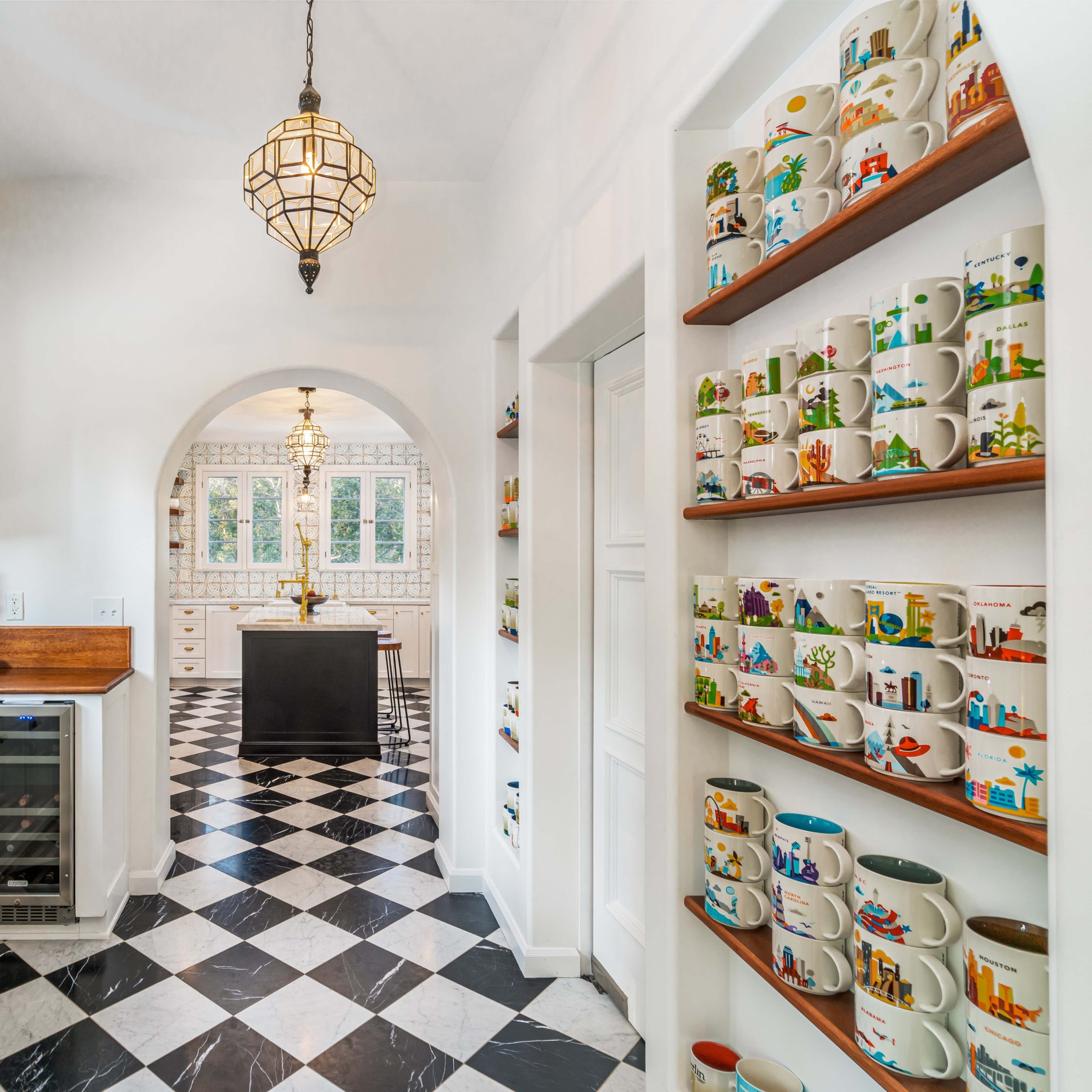 A bright kitchen features white cabinetry, a black and white checkered floor, and shelves lined with colorful mugs.