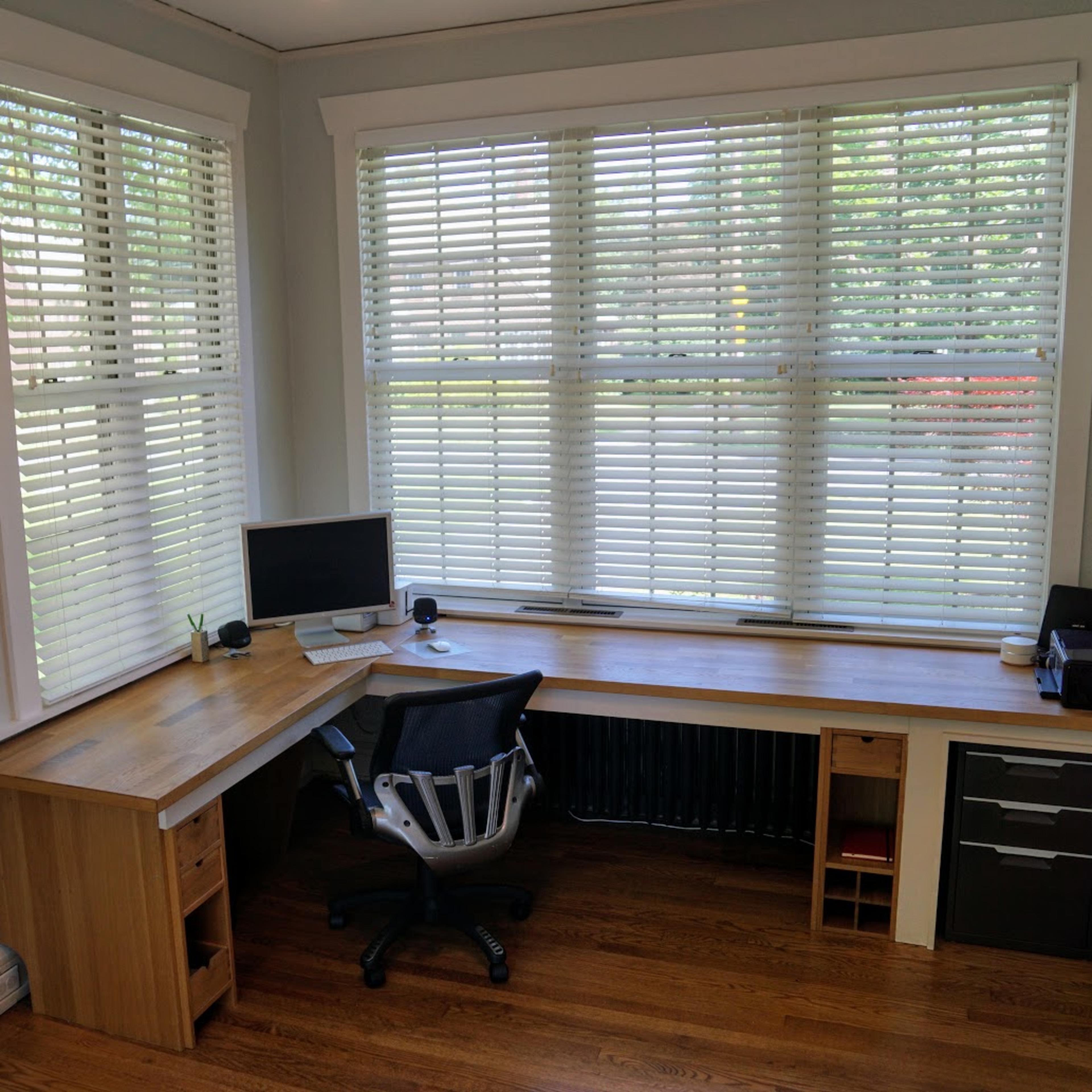 A corner office space features a wooden desk, a computer monitor, and large windows with blinds, providing natural light.