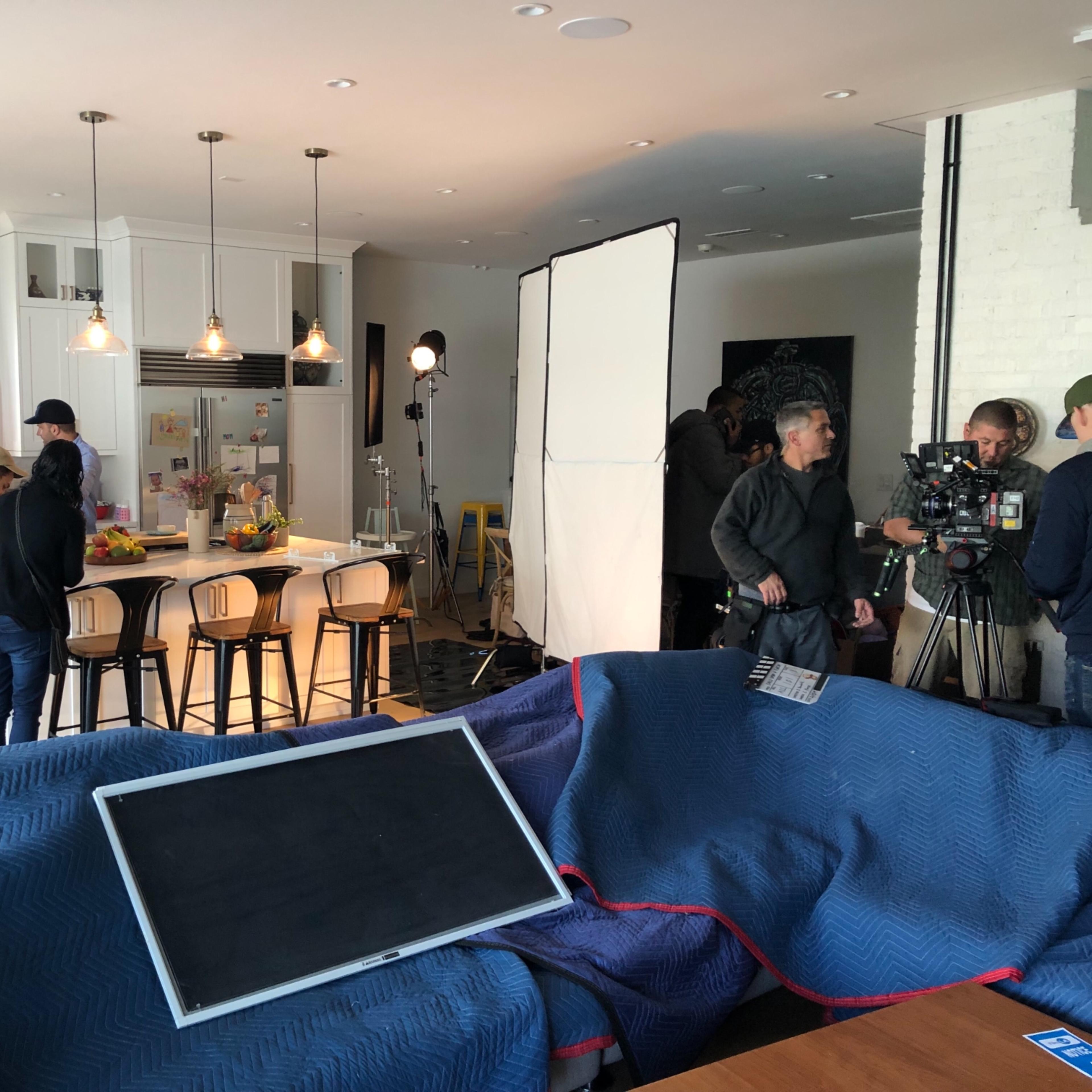 A film crew sets up equipment in a bright kitchen while several people gather around preparing for a shoot.
