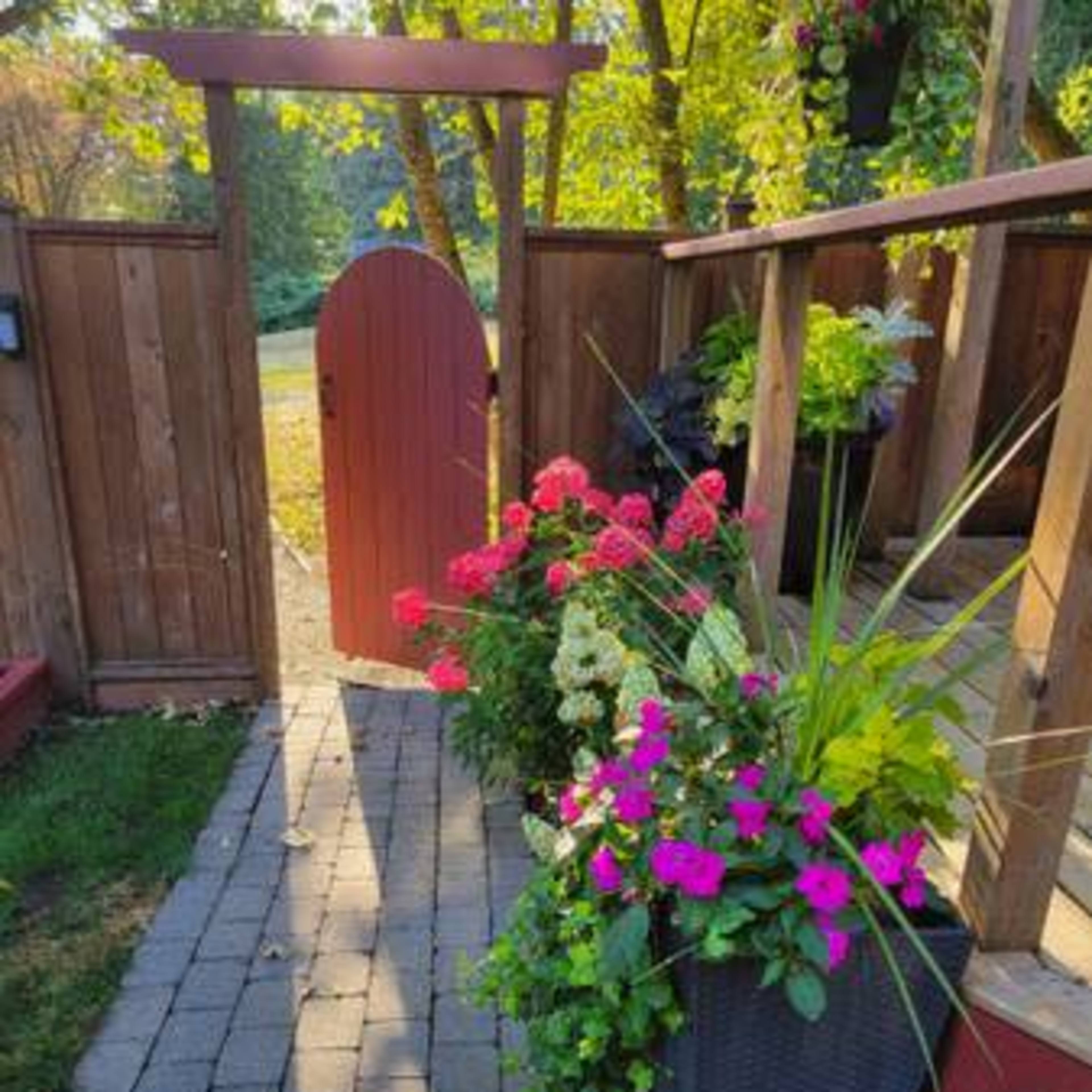 A slate pathway leads through a garden with colorful flower pots and a wooden gate in the background.