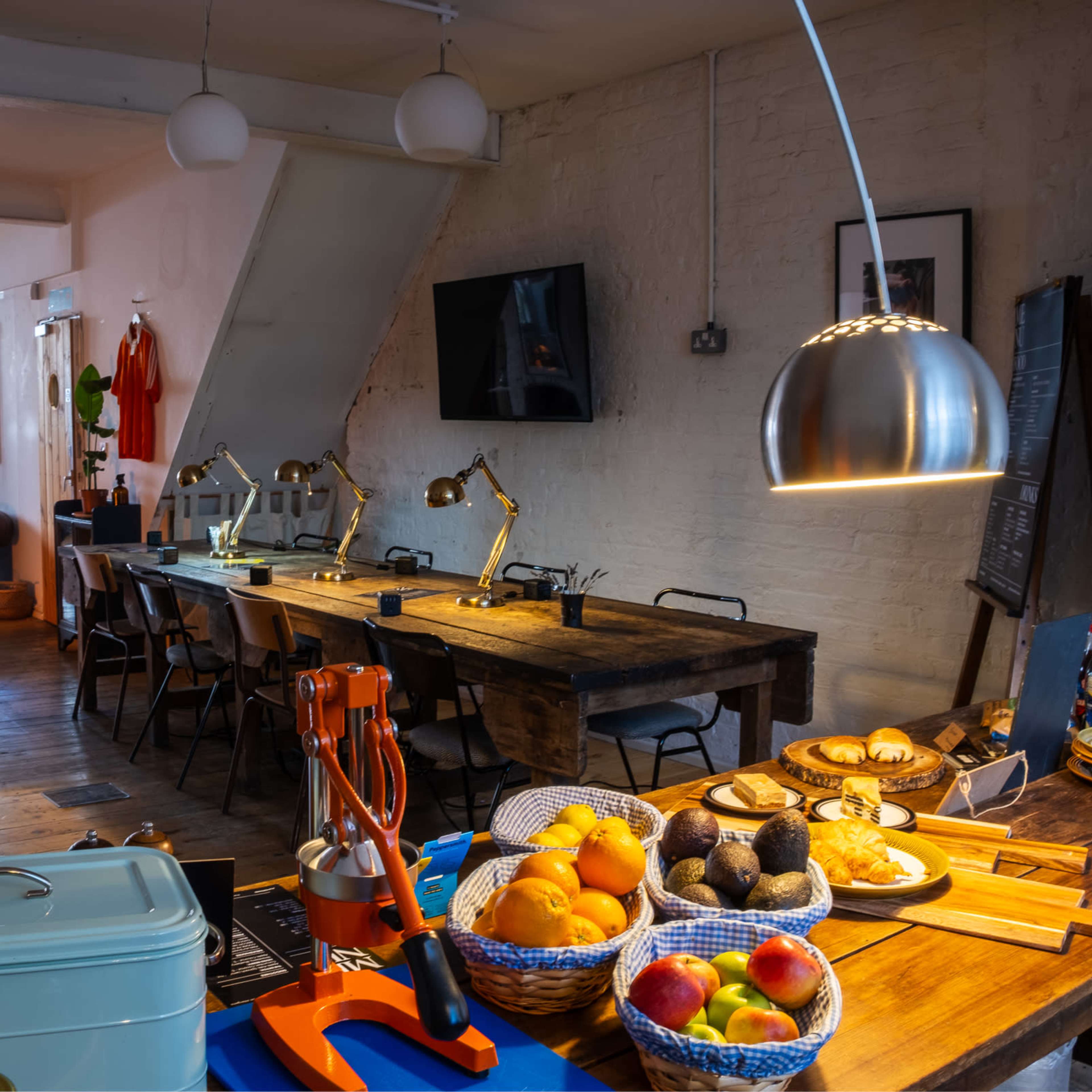 A spacious interior with a kitchen area featuring a table laden with fruits and baked goods, while a living space is visible in the background with a sofa and workspace.
