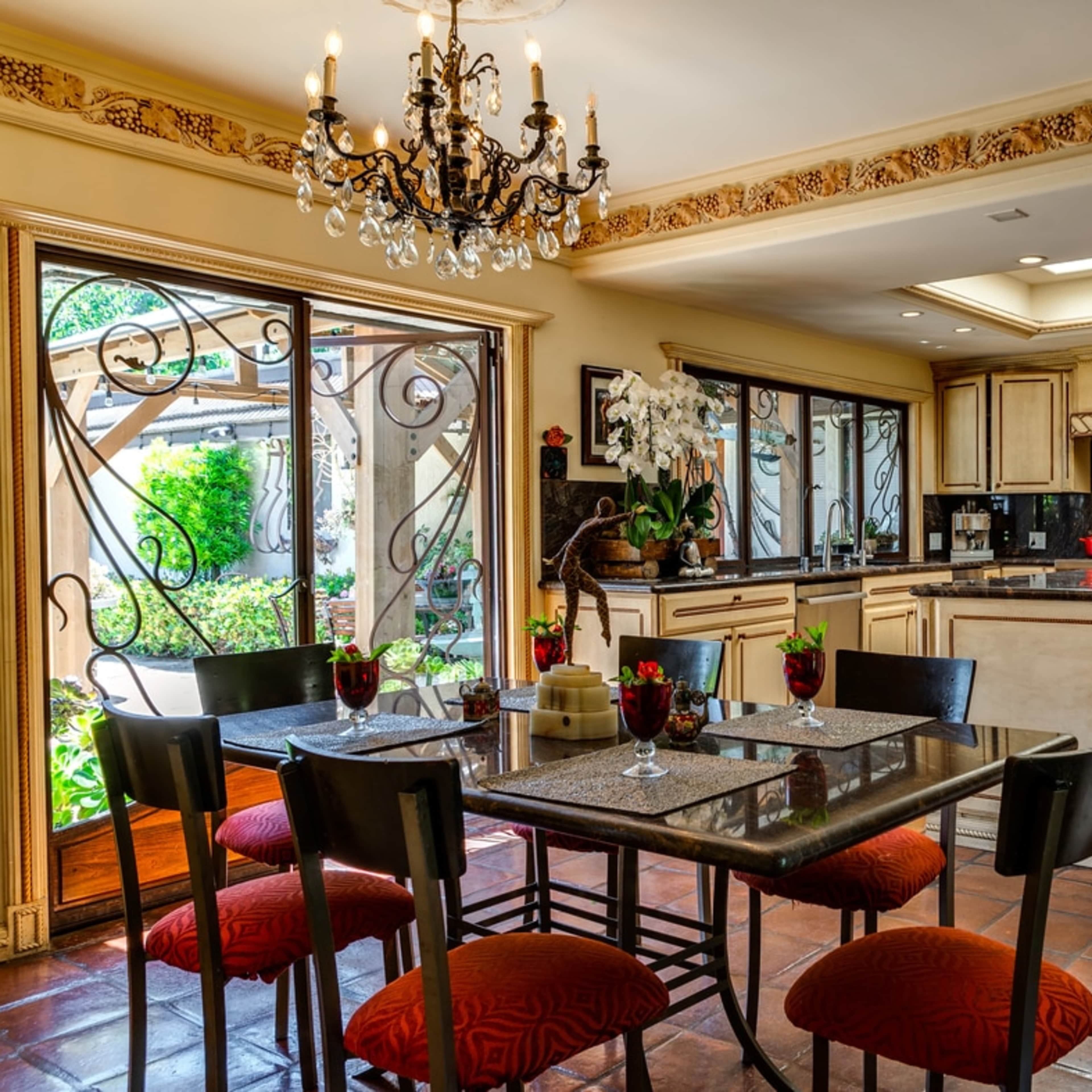 A dining area with a table set for six, featuring a chandelier, surrounded by large windows and a view of a garden, complemented by a kitchen with light-colored cabinetry.