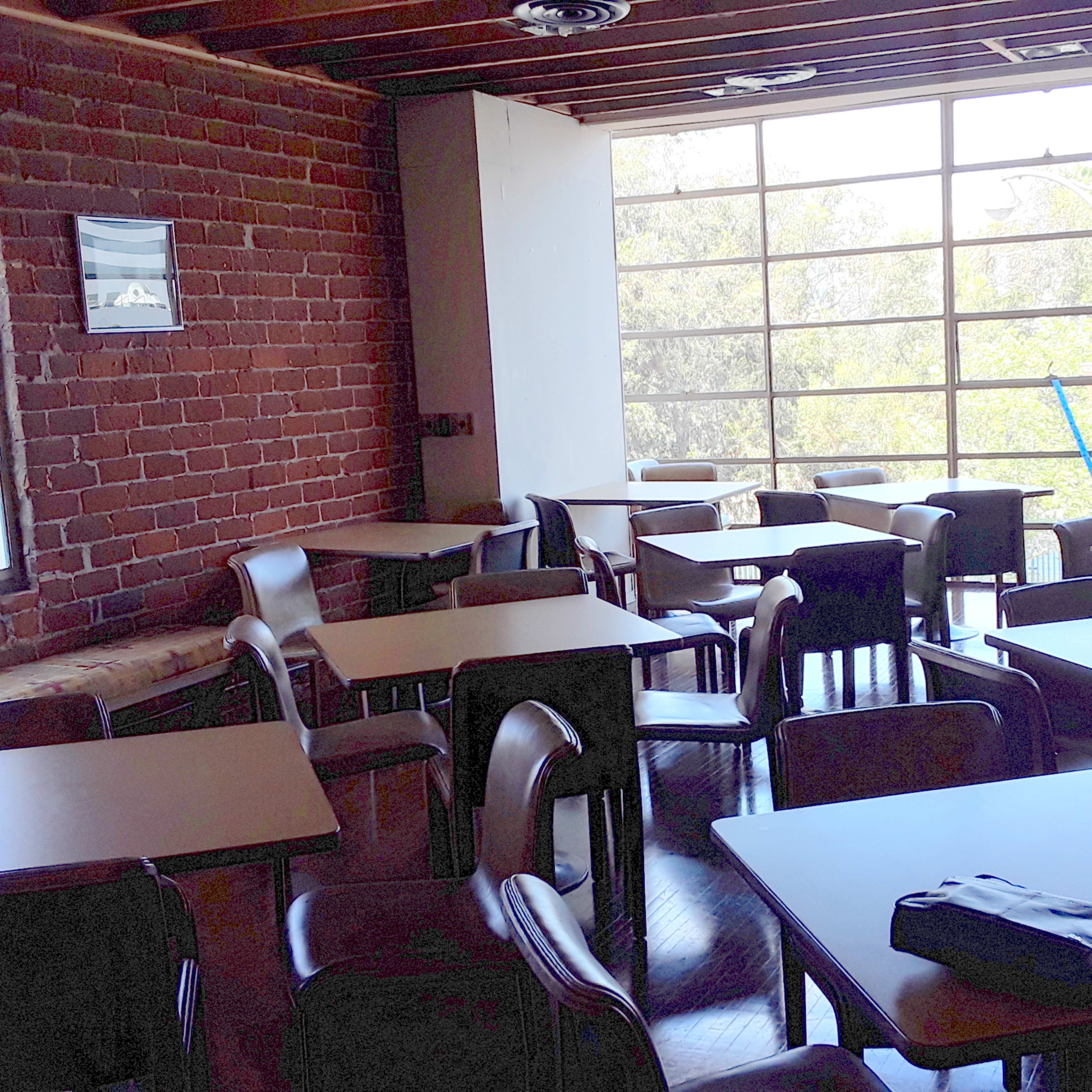 A dining area featuring several empty tables and chairs arranged in a spacious room with large windows and a brick wall.