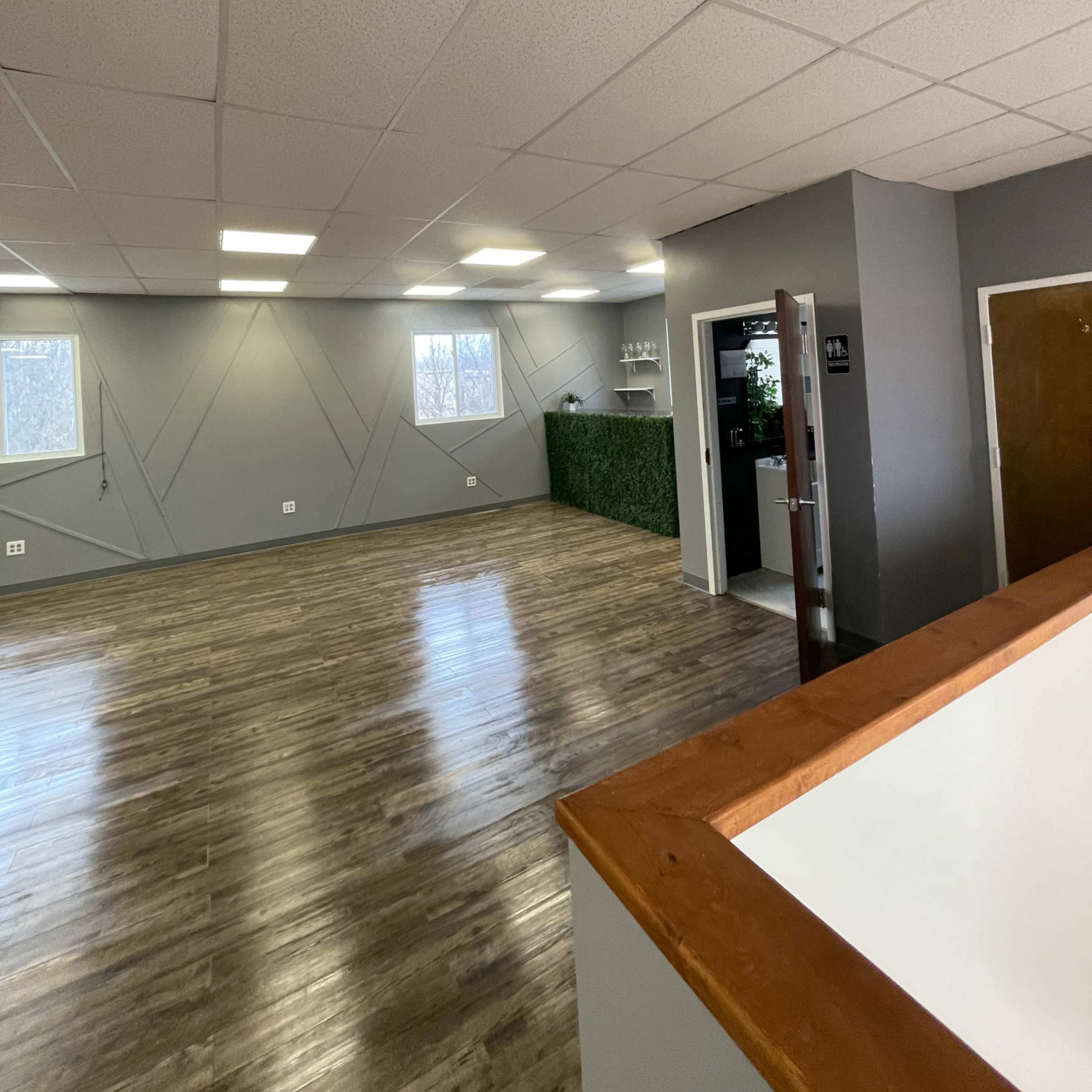 An interior room with light gray walls, wooden flooring, and a green accent wall, featuring windows and an entrance to another area.