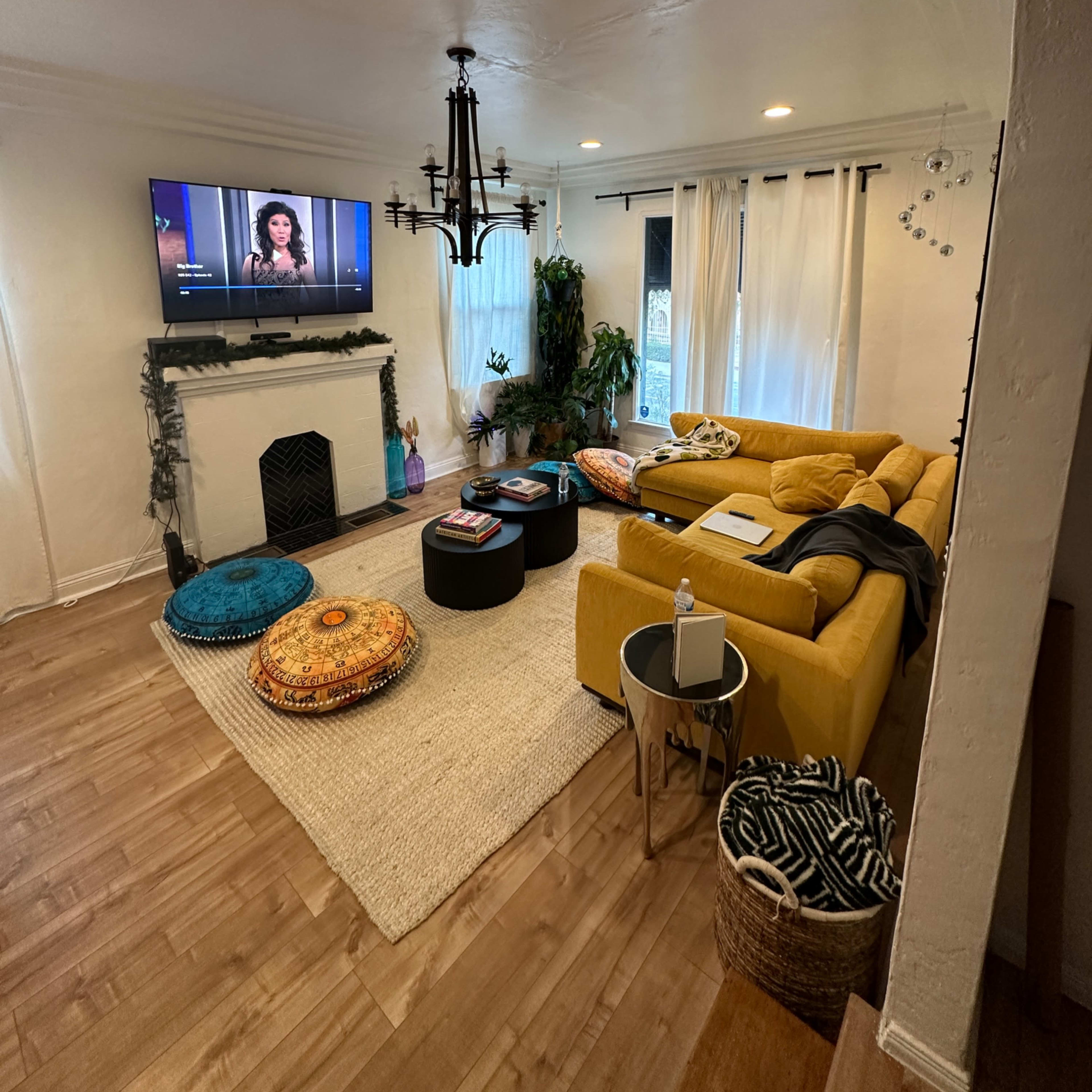 A living room with a yellow sectional sofa, a television mounted on the wall, a fireplace, and various decorative elements including plants and poufs.