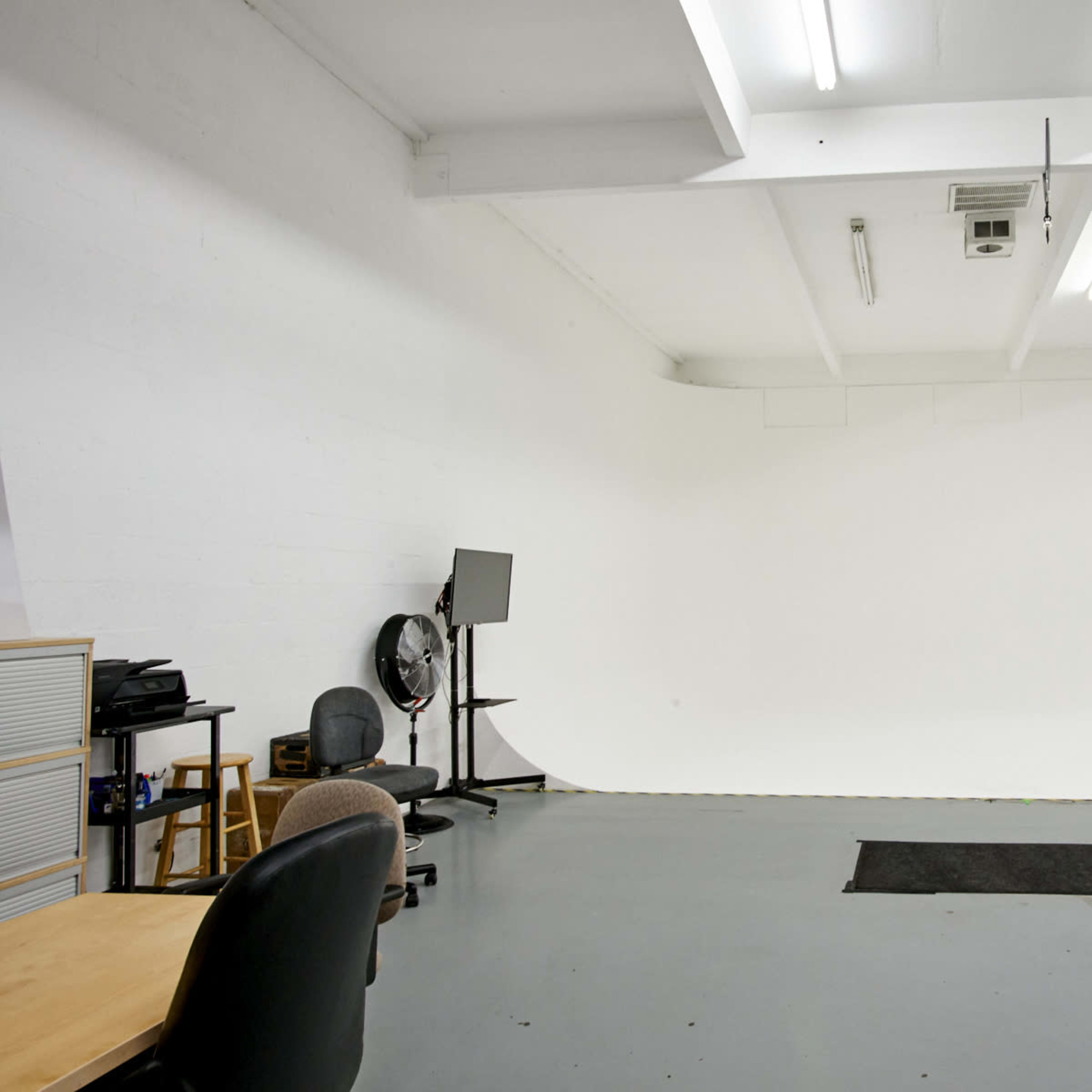 A spacious, empty studio with a white backdrop, gray floor, and minimal furnishings including chairs and a desk.
