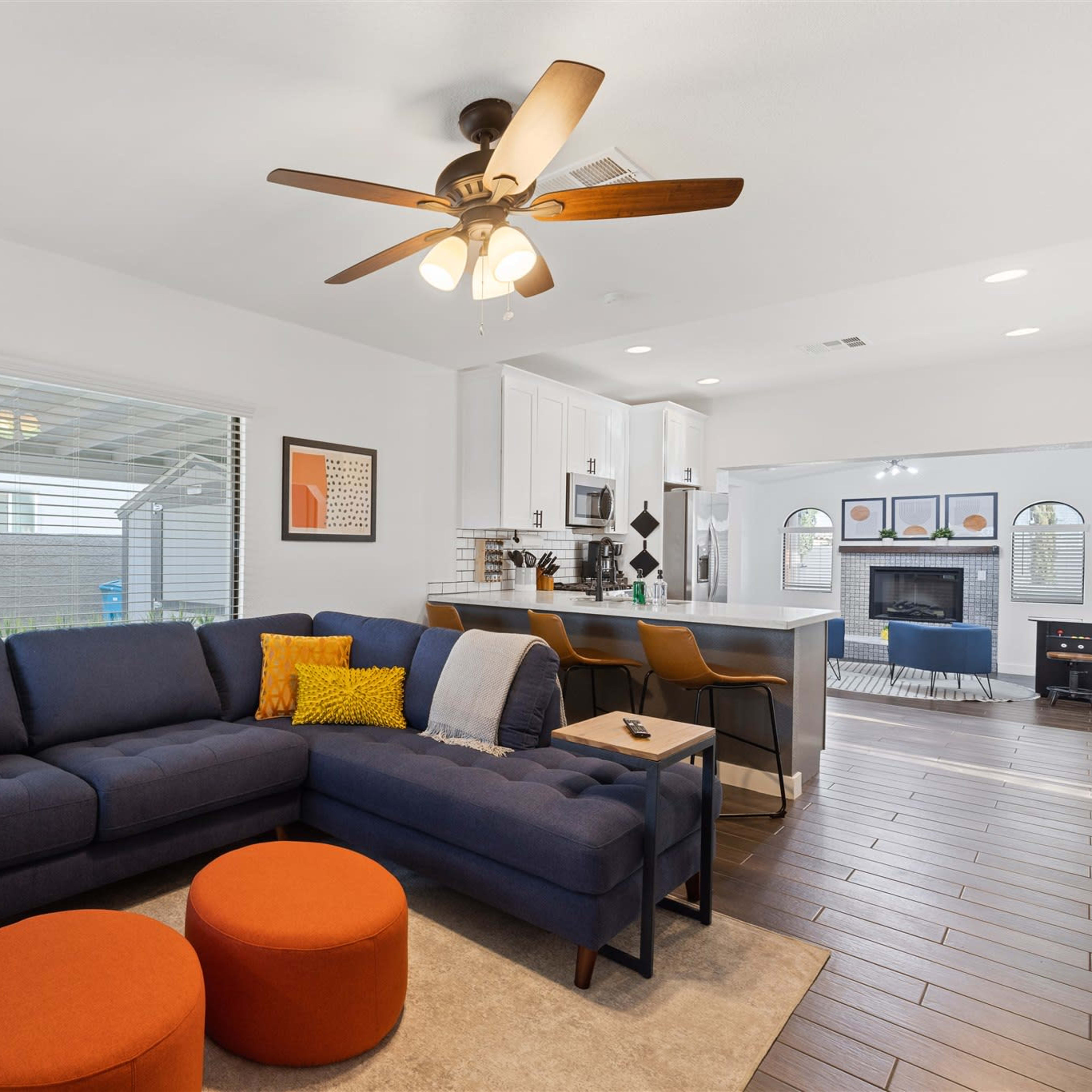 A modern living room features a blue sectional sofa with orange accent pillows and two round orange ottomans, opening to a kitchen with white cabinetry and a view of the backyard.