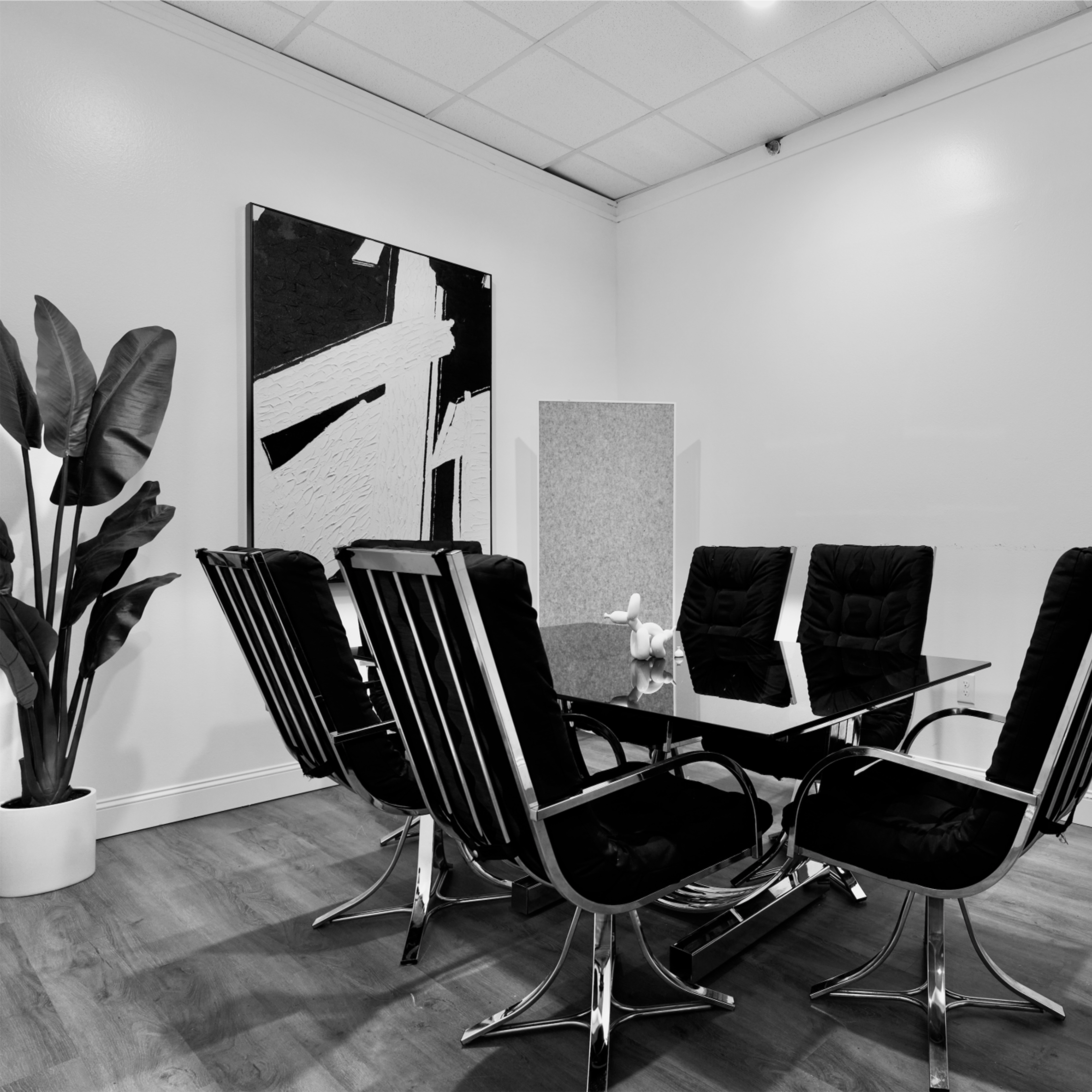 A modern conference room features a glass table surrounded by six black chairs, with large indoor plants in white pots on either side.